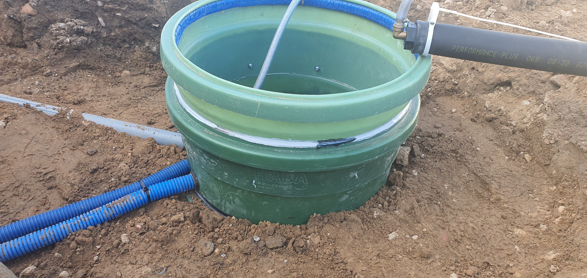 Zielony zbiornik z tworzywa sztucznego, prawdopodobnie część instalacji sanitarnej, umieszczony w wykopie ziemnym, z podłączonymi niebieskimi rurami karbowanymi i czarną rurą Performance Plus...