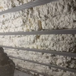 Poddasze z widocznymi metalowymi profilami wypełnione białą, gęstą pianą izolacyjną o nieregularnej strukturze.