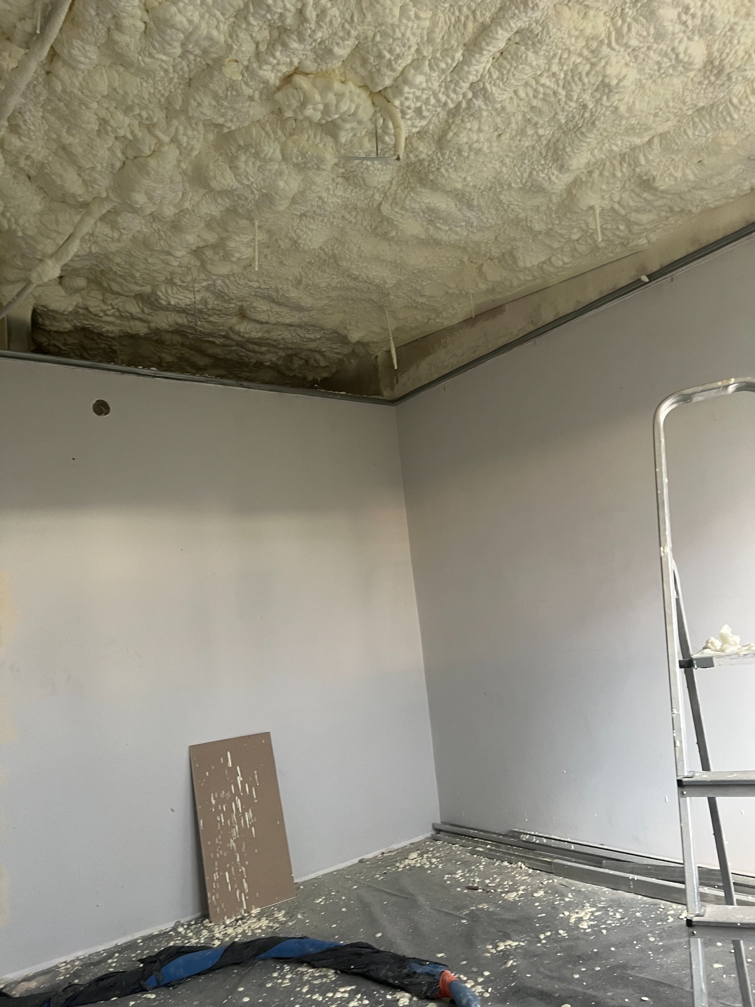 Poddasze po aplikacji izolacji pianką PUR, widoczne nierównomierne pokrycie sufitu pianą, resztki piany na podłodze zabezpieczonej folią, drabina oparta o ścianę.