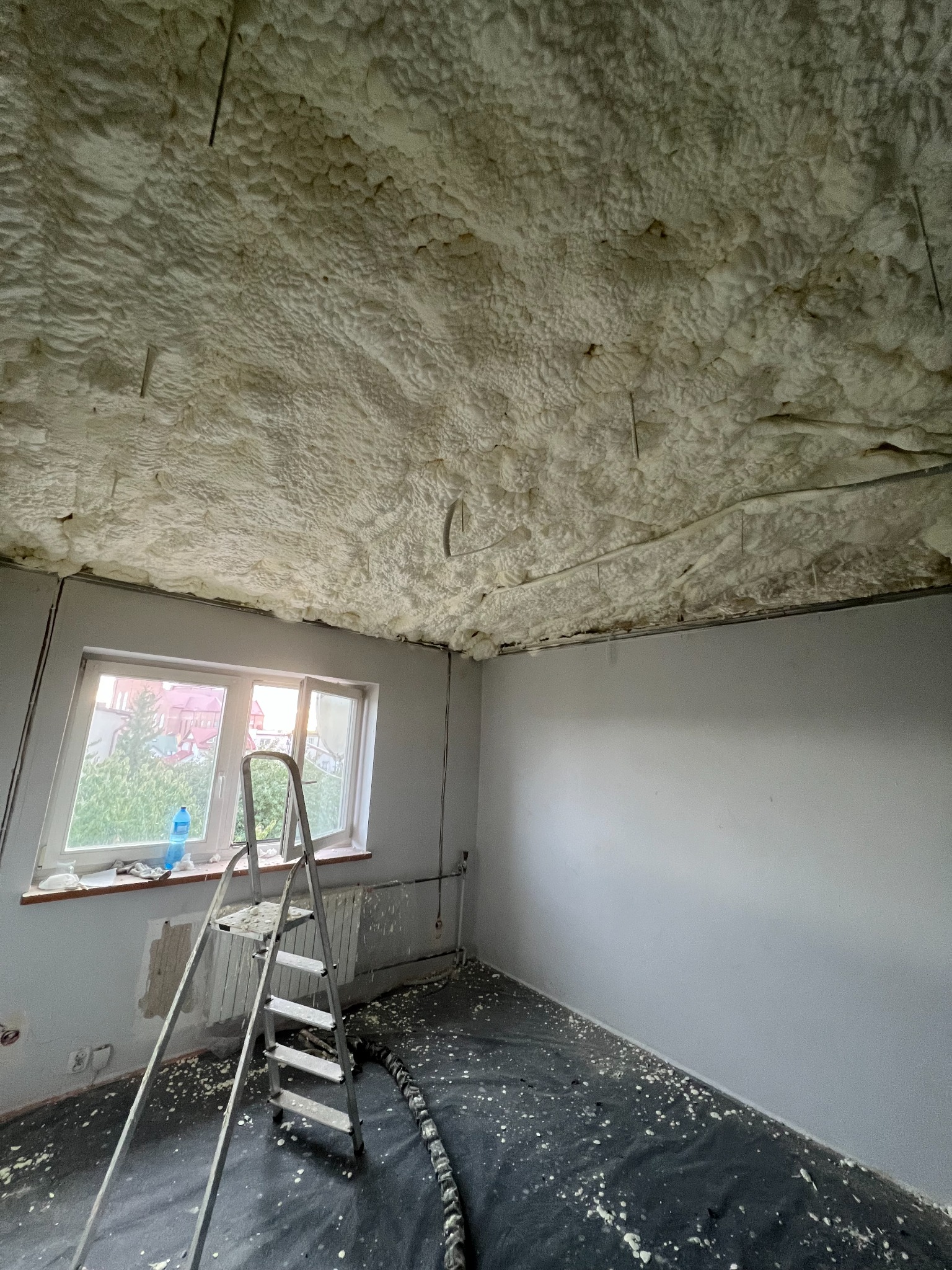 Poddasze po aplikacji szarej piany izolacyjnej, widoczna drabina oparta o okno, podłoga zabezpieczona czarną folią, resztki piany na folii i parapecie.