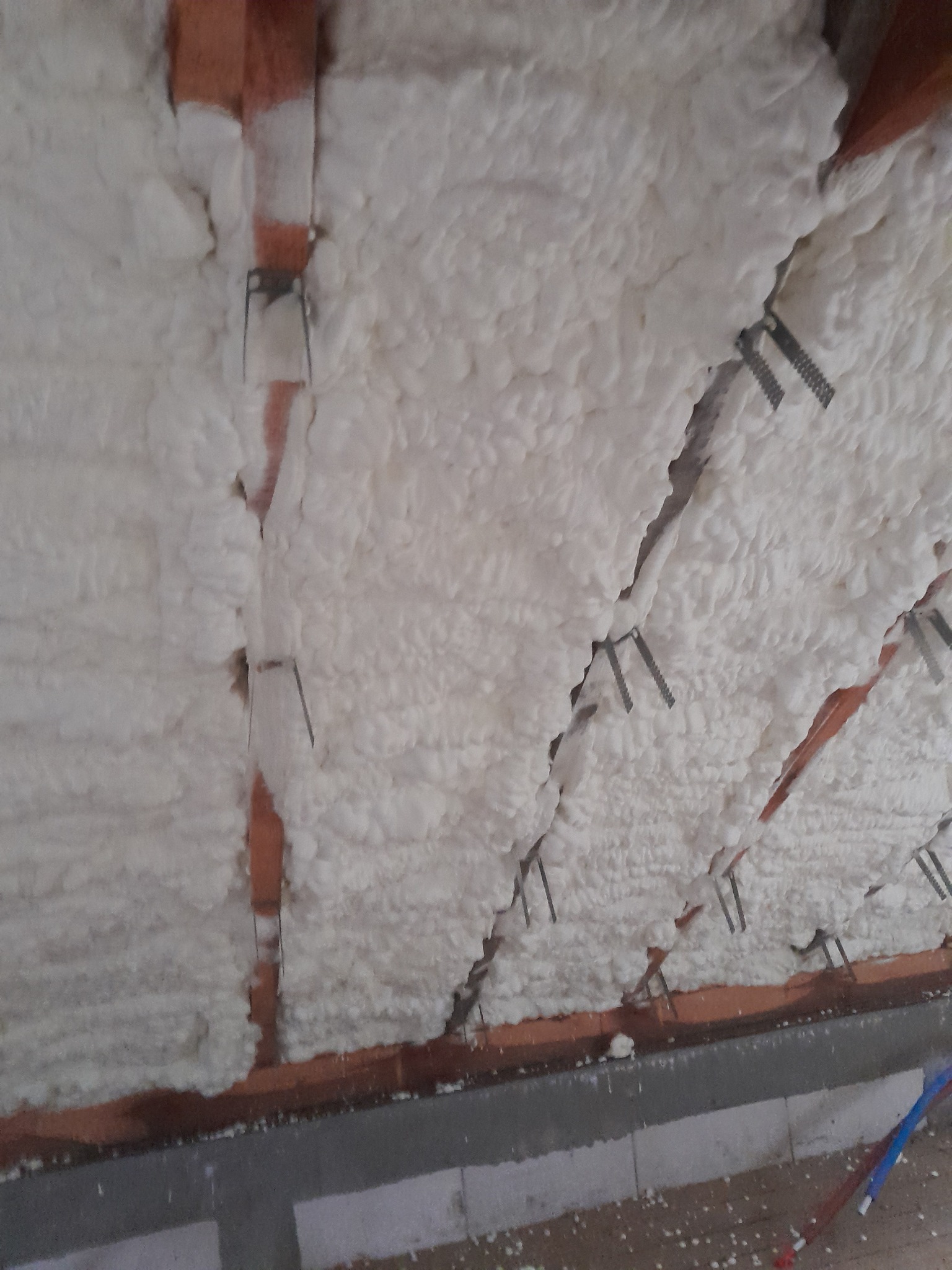 Poddasze z widoczną białą pianką izolacyjną między krokwiami, mocowaną metalowymi klamrami do drewnianych belek.