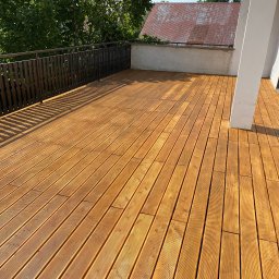 InSteel&Glass Miłosz Cieplik - Jasny, słoneczny taras z desek z drewnianą balustradą i widokiem na dach domu oraz zieleń drzew.