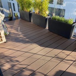 InSteel&Glass Miłosz Cieplik - Balkon z brązową podłogą z desek, czarne donice z bambusem i lawendą, klimatyzator oraz worek ziemi.