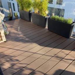 Balkon z brązową podłogą z desek, czarne donice z bambusem i lawendą, klimatyzator oraz worek ziemi.