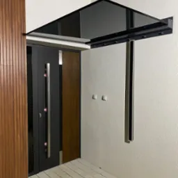 Nowoczesne wejście do domu z czarną, szklaną osłoną nad drzwiami, wspartą na metalowych elementach, obok drewniana okładzina ściany i jasna kostka brukowa.