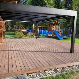 InSteel&Glass Miłosz Cieplik - Drewniany taras z czarną pergolą metalową, z widokiem na plac zabaw z basenem i domkiem. Deski tarasowe ułożone równolegle, z widoczną strukturą drewna.