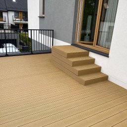 InSteel&Glass Miłosz Cieplik - Nowoczesny taras kompozytowy z czarną balustradą i schodami, z widokiem na dom. Jasne, naturalne oświetlenie podkreśla detale desek tarasowych.