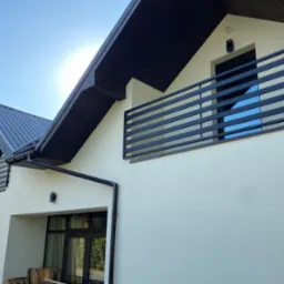 Nowoczesny dom z beżową elewacją, czarną balustradą balkonową z poziomych profili i ciemnym dachem. Widok z dołu, błękitne niebo w tle.