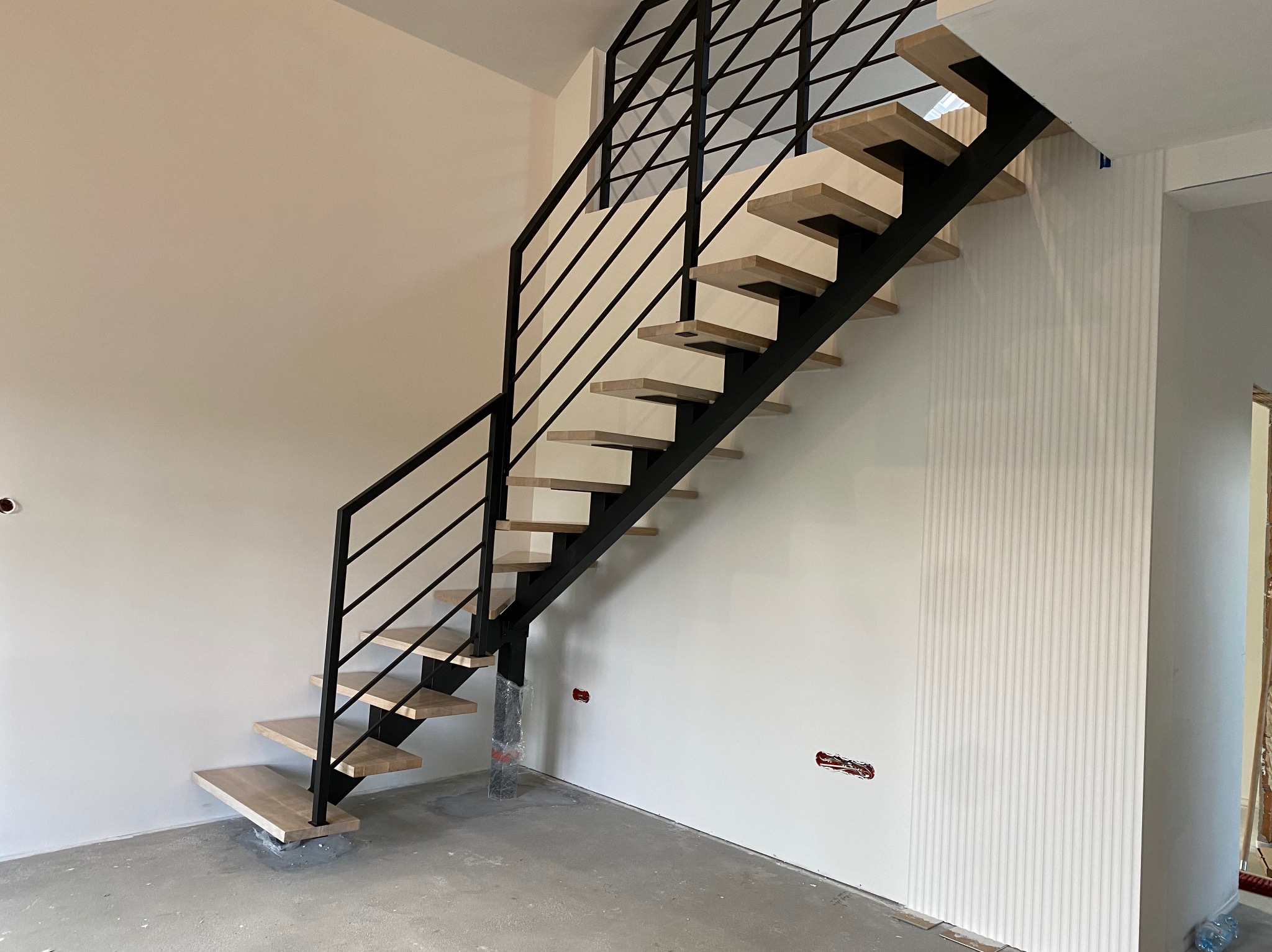 Nowoczesne schody metalowe z drewnianymi stopniami, czarna balustrada. Minimalistyczny design, surowe wnętrze w trakcie wykańczania. Widoczne punkty elektryczne na ścianie.