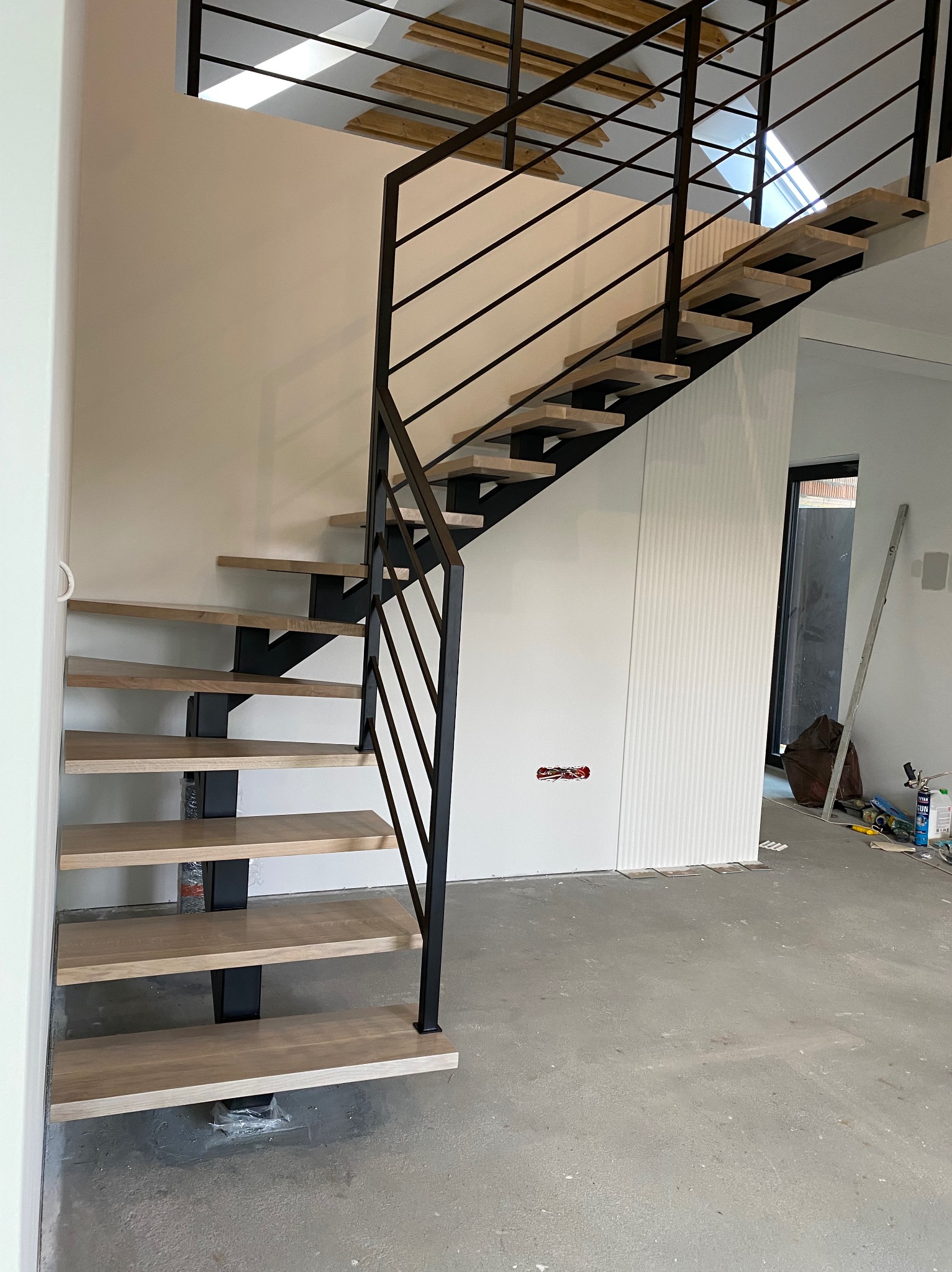 Nowoczesne schody metalowe z drewnianymi stopniami, czarna balustrada, surowe wnętrze w trakcie wykończenia, widoczne narzędzia i materiały budowlane.