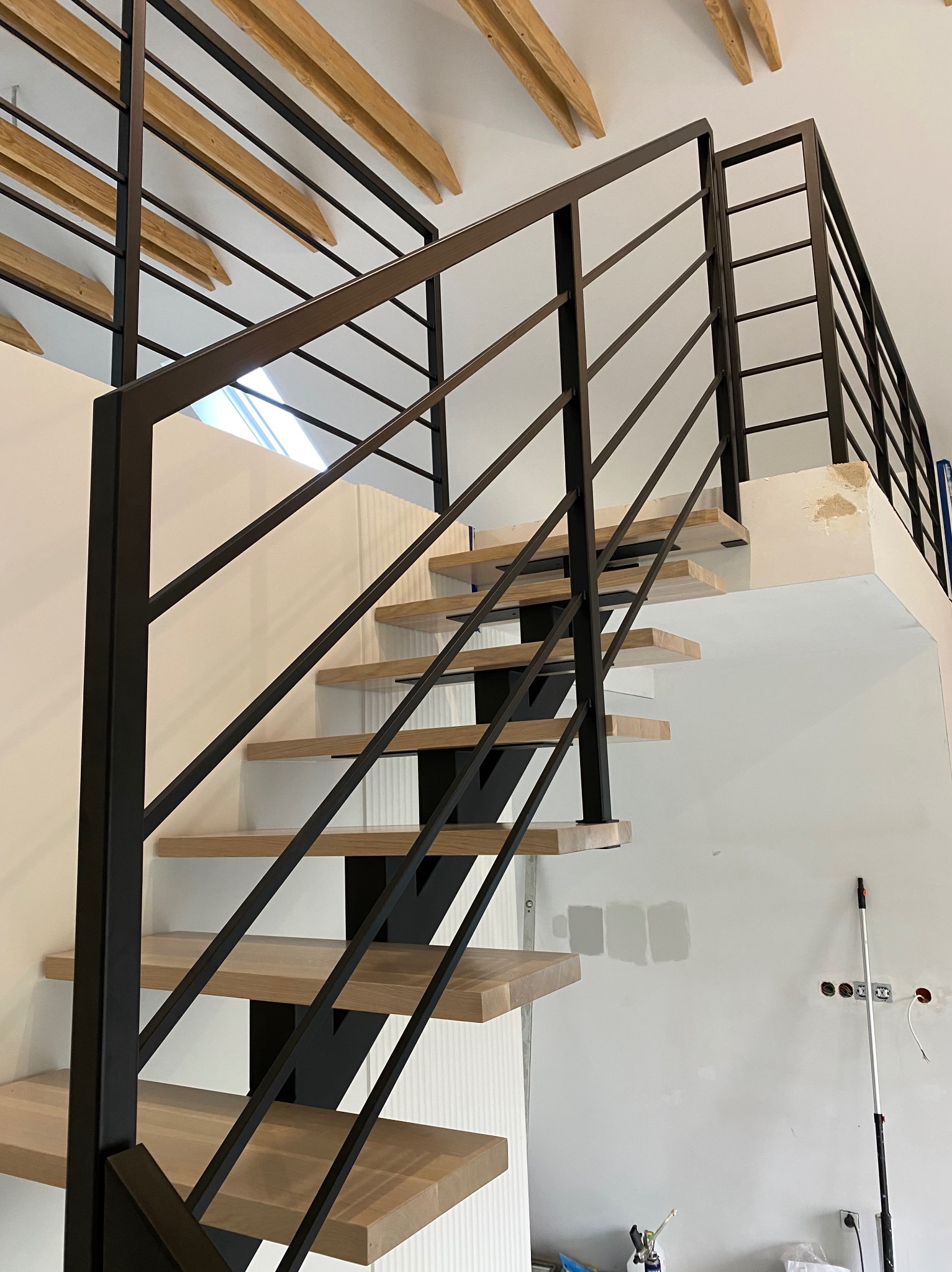 Nowoczesne schody metalowe z drewnianymi stopniami, czarna balustrada z poziomymi prętami, surowe wykończenie wnętrza w tle. Widok z dołu.