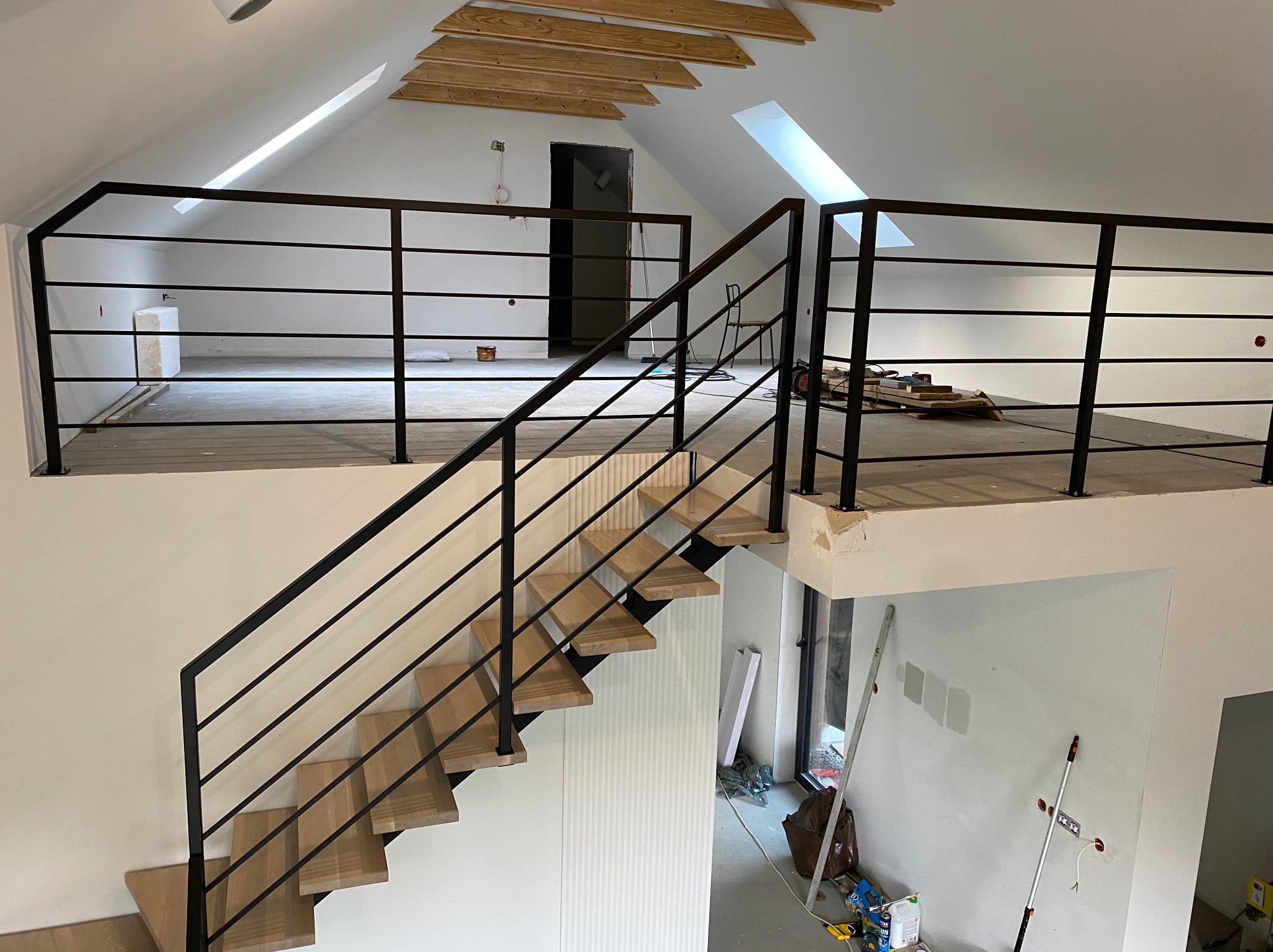Nowoczesne schody drewniane z czarną, metalową balustradą w jasnym wnętrzu domu z poddaszem. Widoczne okna dachowe i elementy wykończeniowe.