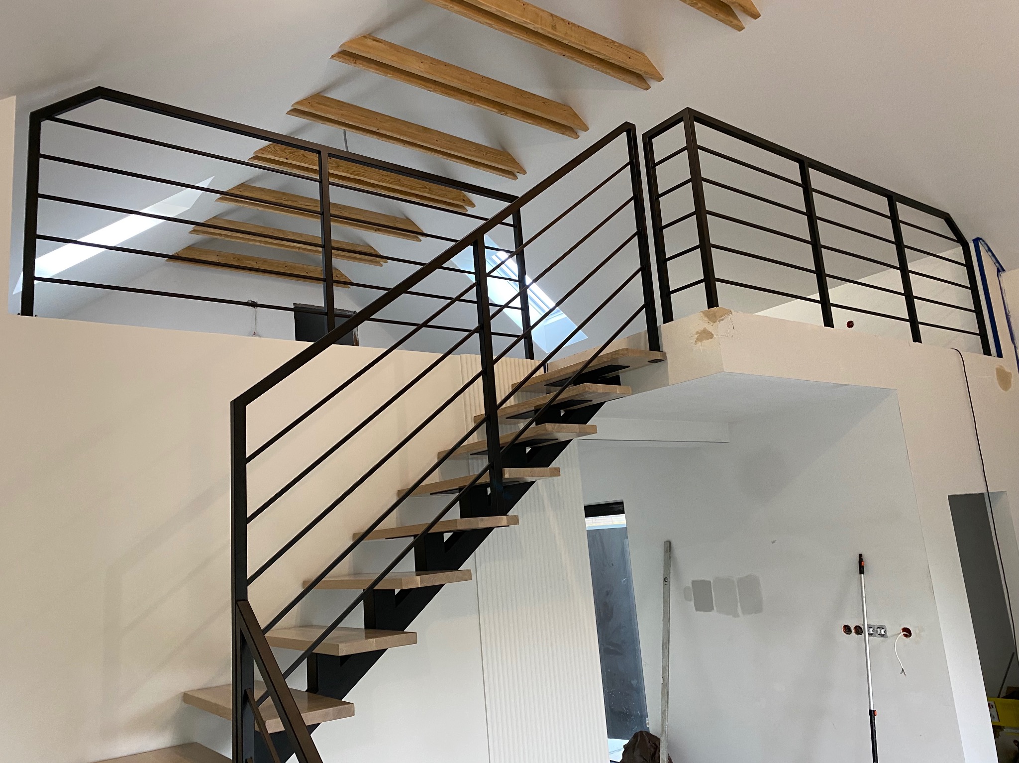 Nowoczesne schody z czarną metalową balustradą i drewnianymi stopniami w jasnym wnętrzu z drewnianymi belkami stropowymi na poddaszu.