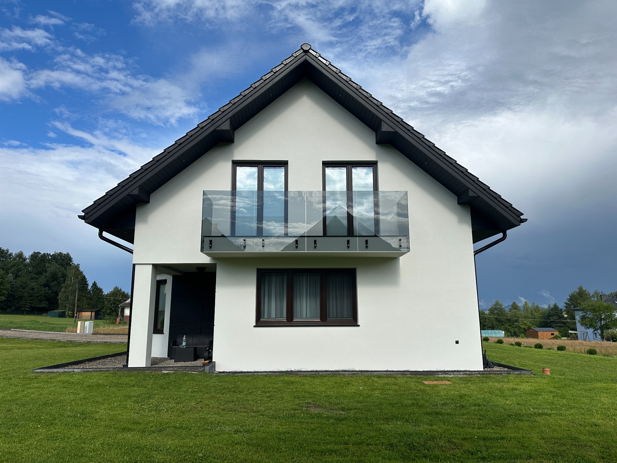 Biały dom z balkonem zabezpieczonym szkłem, odbijającym błękitne niebo. Ciemna stolarka okienna i dach kontrastują z jasną elewacją. Soczysta zieleń trawnika.