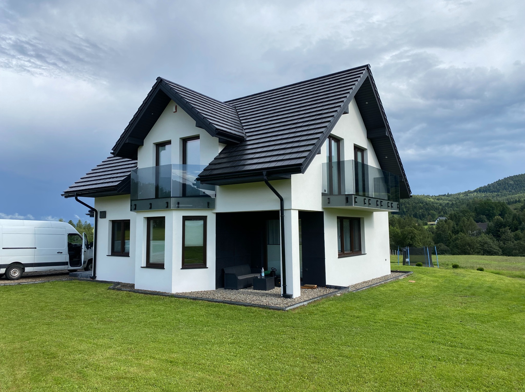 Nowoczesny dom z białymi ścianami i czarnym dachem. Balkony zabezpieczone szkłem. Widoczny fragment vana.