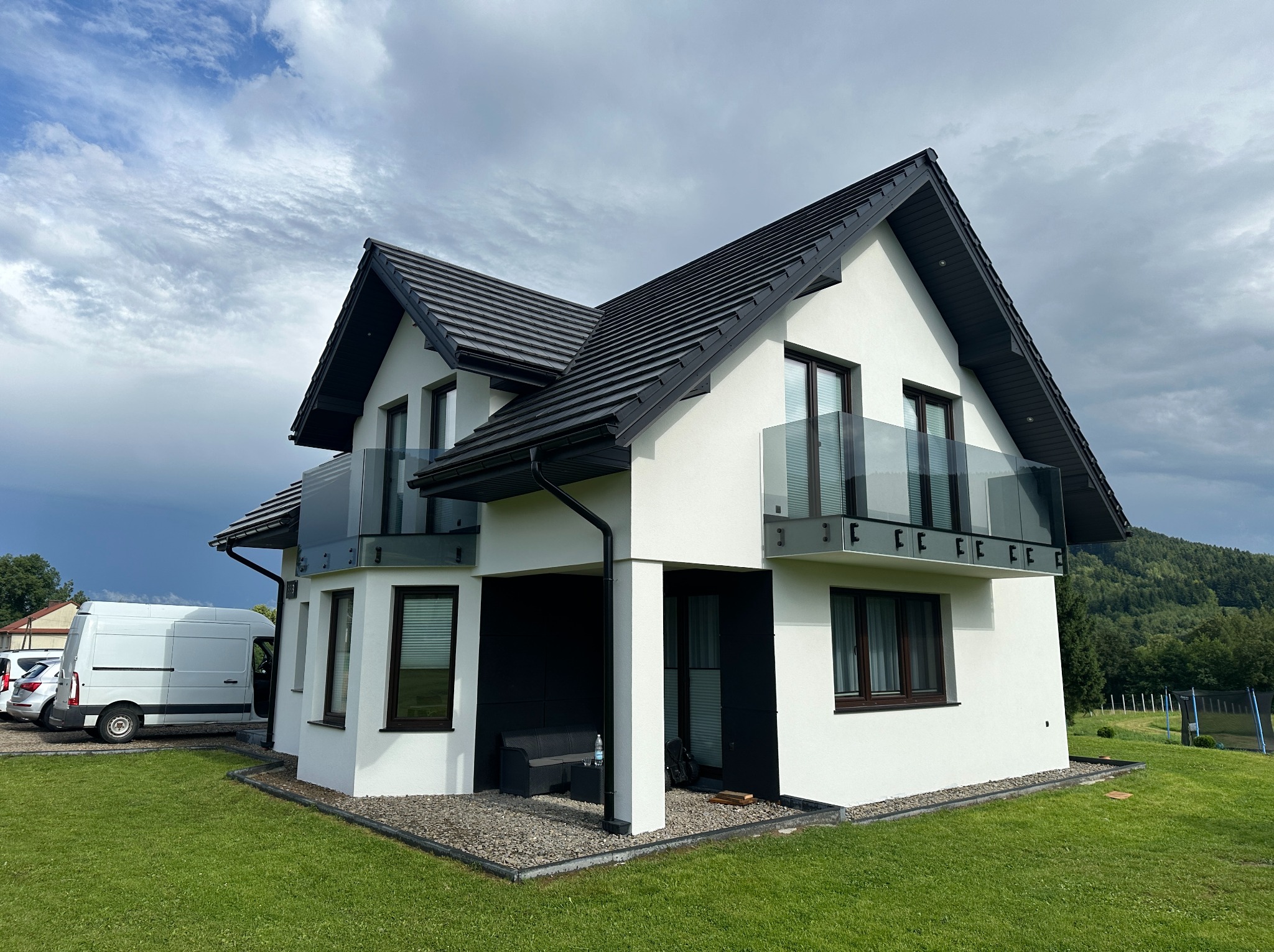 Nowoczesny dom z białymi ścianami i czarnym dachem. Szklane balustrady balkonowe, zieleń trawnika i błękitne niebo w tle. Widok z zewnątrz, dzień.