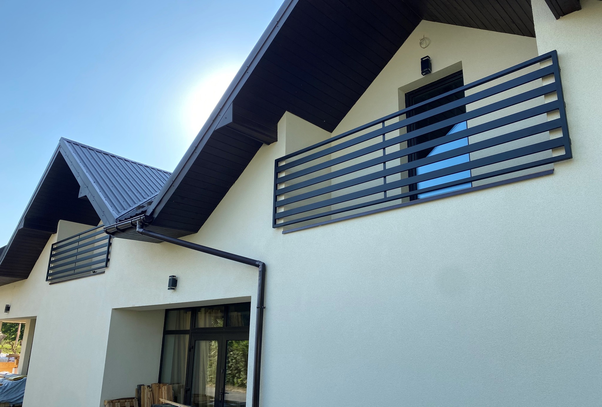 Nowoczesny dom z beżową elewacją, czarną balustradą balkonową z poziomych profili i ciemnym dachem. Widok z dołu, błękitne niebo w tle.