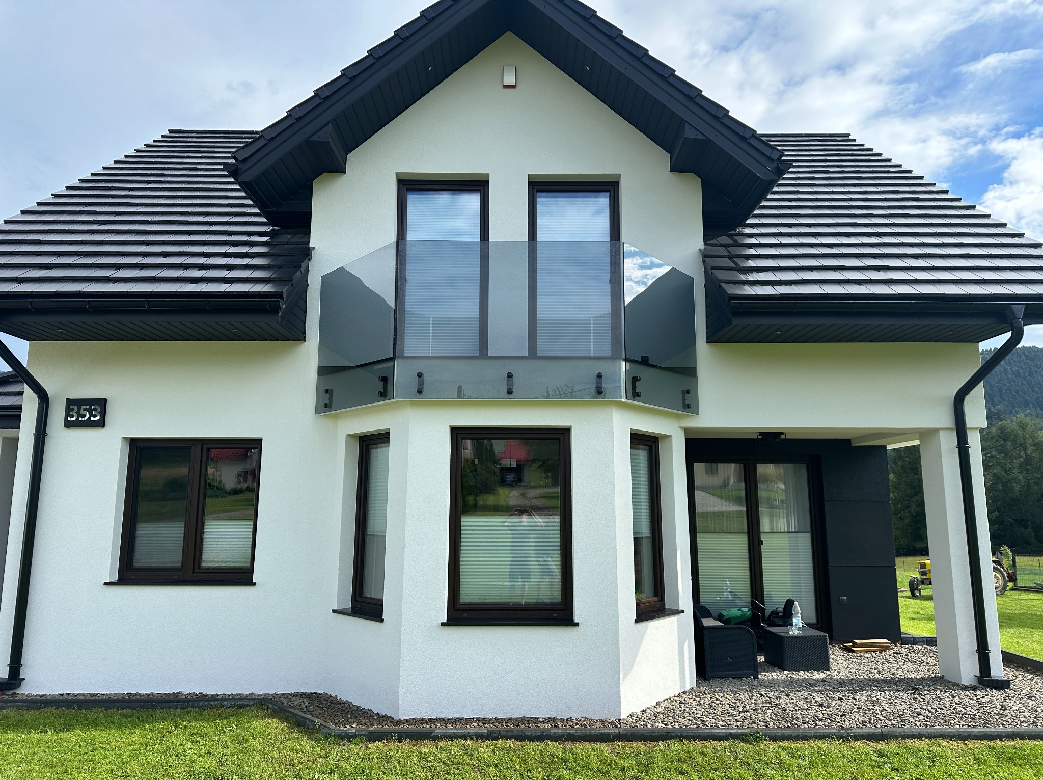 Nowoczesny dom z balkonem zabezpieczonym szklaną balustradą. Widoczne ciemne ramy okienne i czarny dach. Na trawniku stoi żółty traktor.