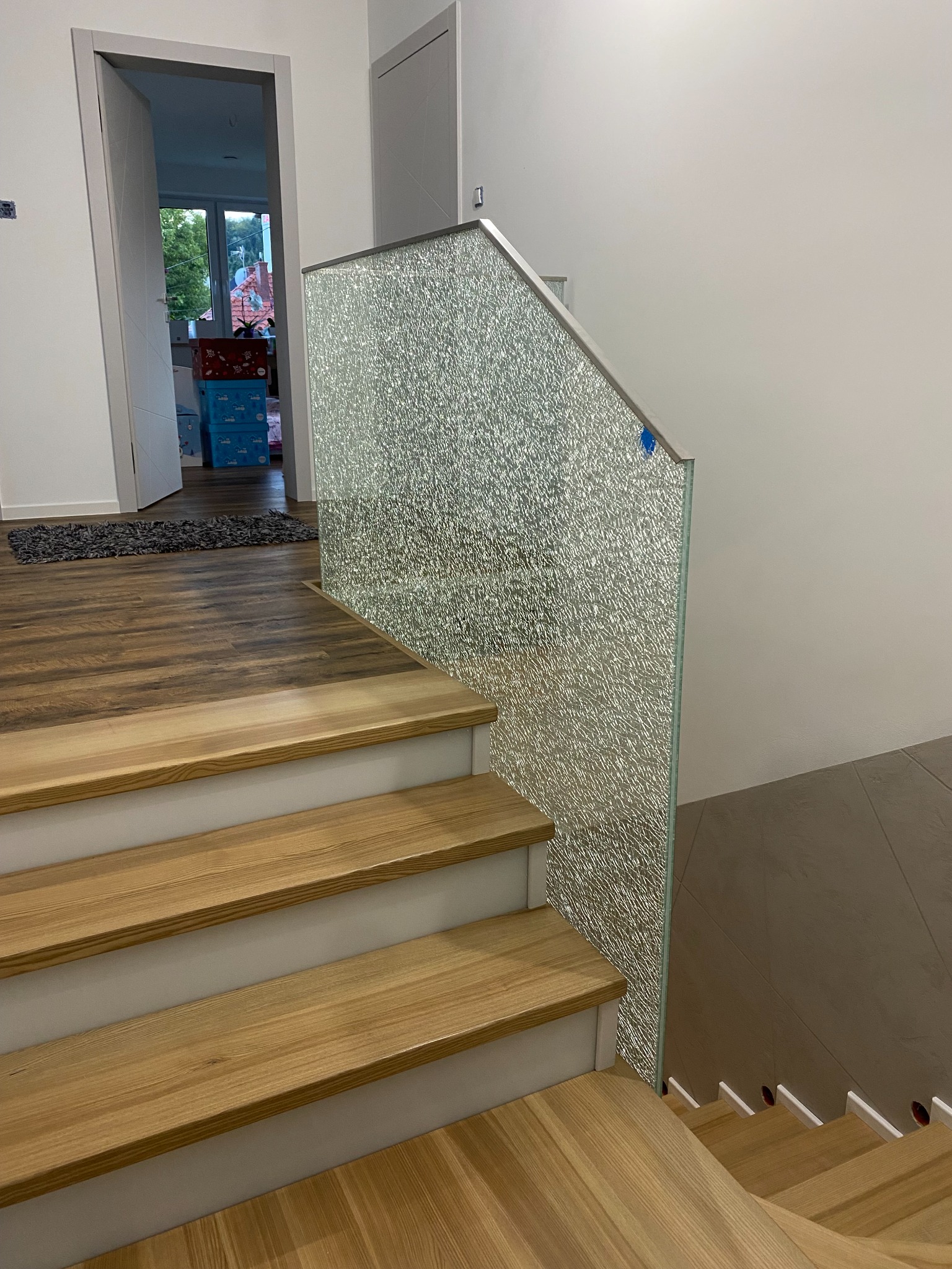 Szklana balustrada z metalowym wykończeniem, zamontowana przy drewnianych schodach wewnątrz jasnego pomieszczenia. Widok z dołu schodów, na pierwszym planie drewniane stopnie.