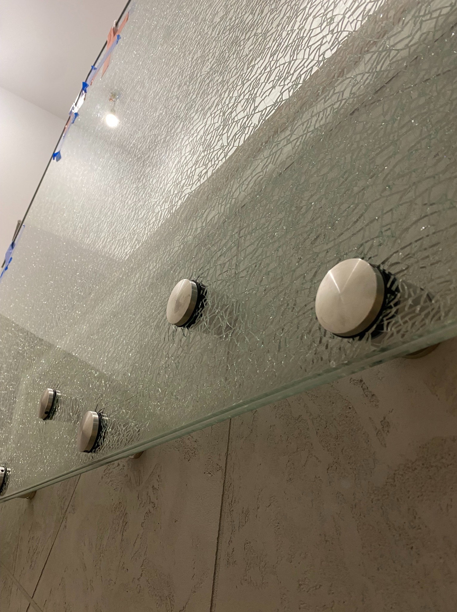 Szklana tafla z dekoracyjną fakturą, zamocowana na metalowych uchwytach. Widoczna struktura ściany w tle.