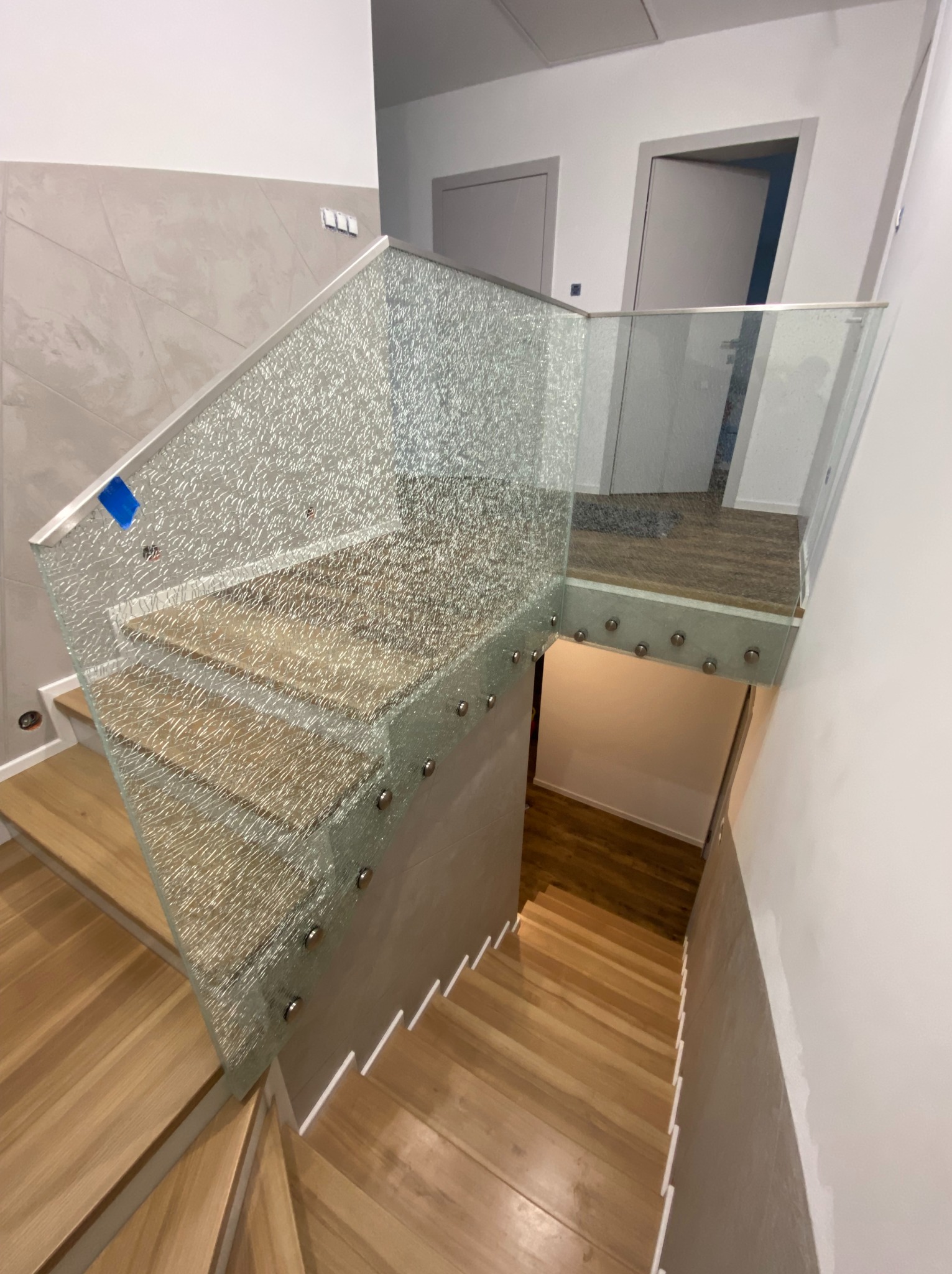 Nowoczesne schody drewniane z balustradą ze szkła ornamentowego, mocowaną punktowo. Widok z góry na dół, minimalistyczny design wnętrza, jasne ściany, drzwi w tle.