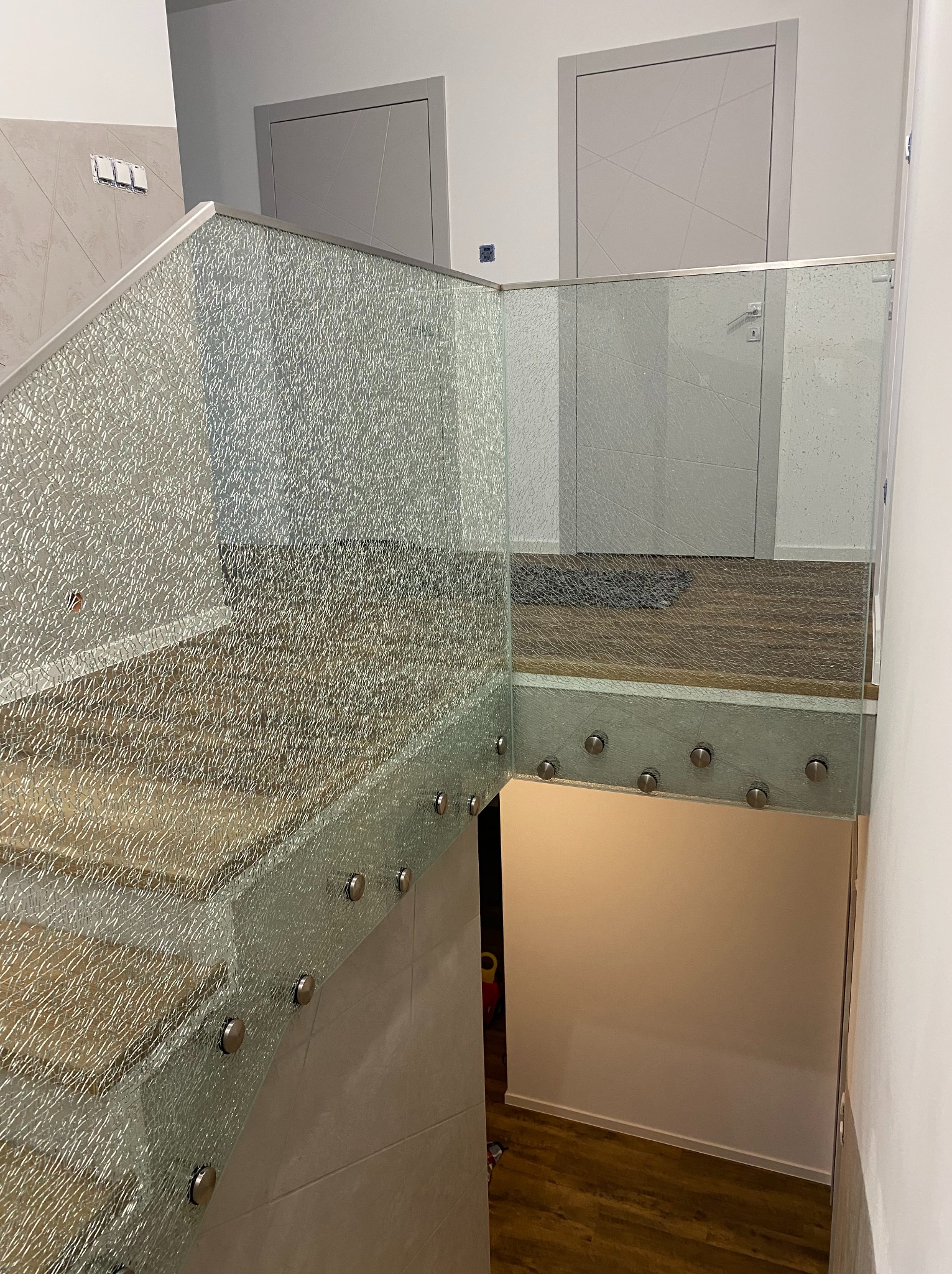 Szklana balustrada schodowa z unikalną fakturą szkła, mocowana punktowo. Widoczne drewniane stopnie i szare drzwi w tle. Nowoczesny design.