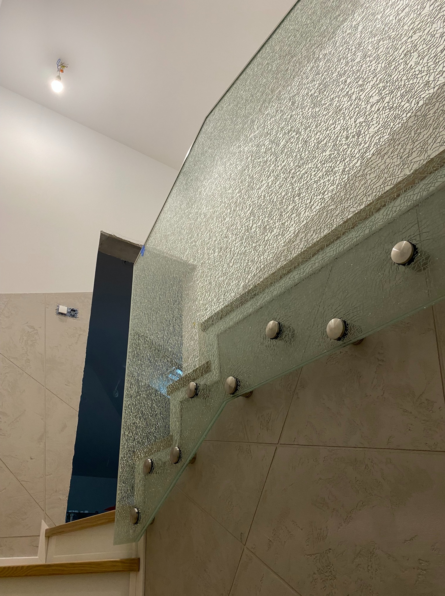 Szklana balustrada schodowa z fakturą, mocowana na metalowych elementach, wnętrze w trakcie wykańczania, widoczne schody i fragment ściany z instalacją elektryczną.