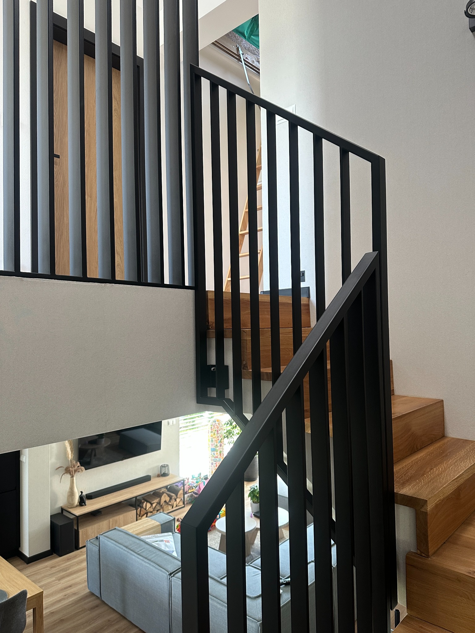 Nowoczesne schody drewniane z czarną, minimalistyczną balustradą metalową, widziane z góry. W tle salon z szarą sofą i drewnianymi elementami wykończenia.