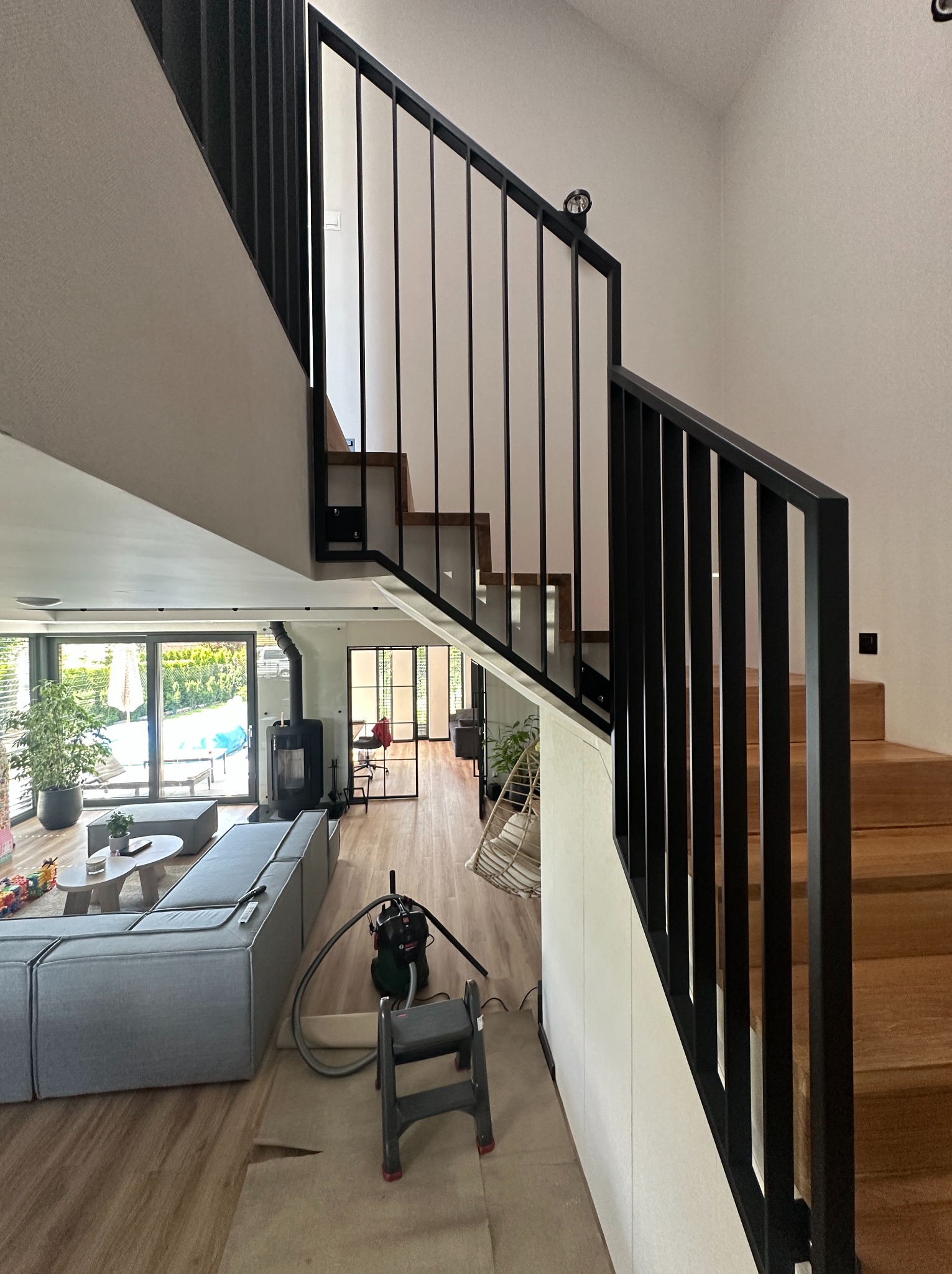 Drewniane schody z czarną, metalową balustradą w nowoczesnym domu. Widok na salon z szarą sofą i kominkiem. W tle ogród za szklanymi drzwiami.