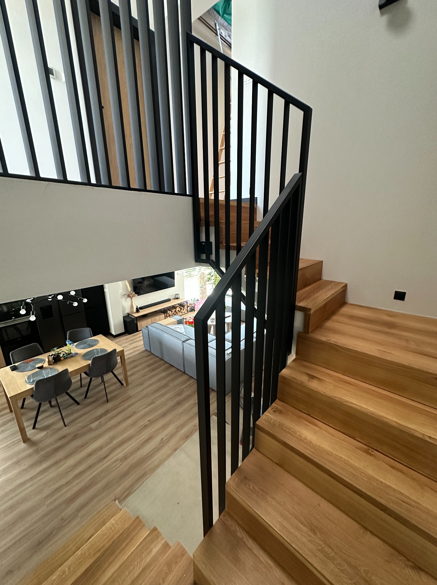 Nowoczesne schody drewniane z czarną metalową balustradą w dwupoziomowym wnętrzu. Widok z góry na salon i jadalnię. Minimalistyczny design i jasne kolory.