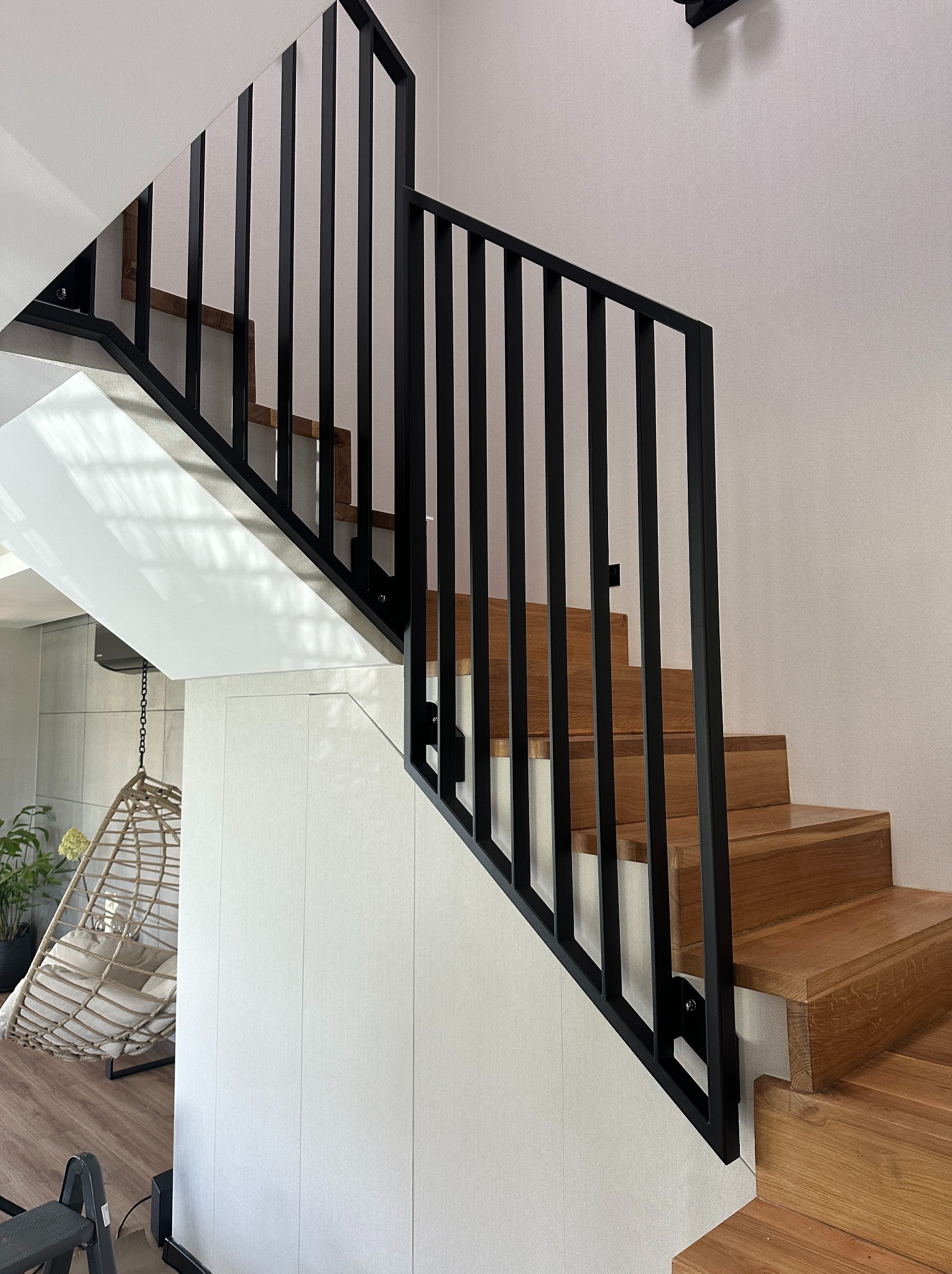 Drewniane schody z czarną metalową balustradą w nowoczesnym wnętrzu. Widoczne stopnie i pionowe elementy balustrady. W tle fotel wiszący.