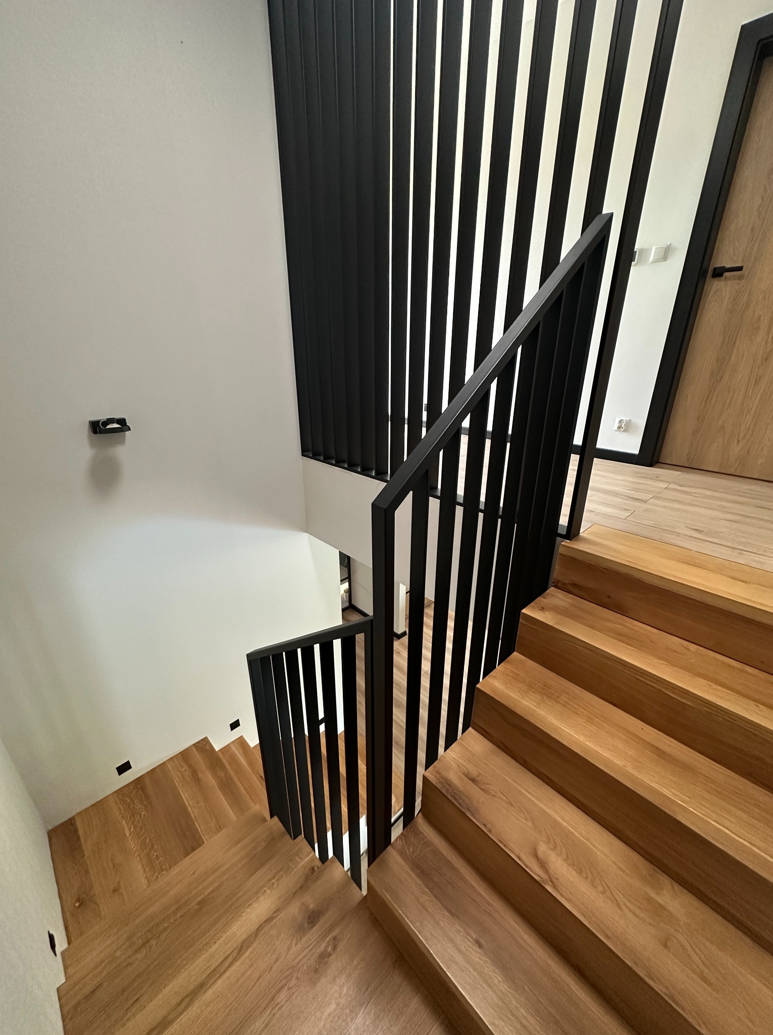 Nowoczesne schody drewniane z czarną, metalową balustradą i pionowymi elementami dekoracyjnymi, widok z góry. Minimalistyczne oświetlenie punktowe wzdłuż schodów.