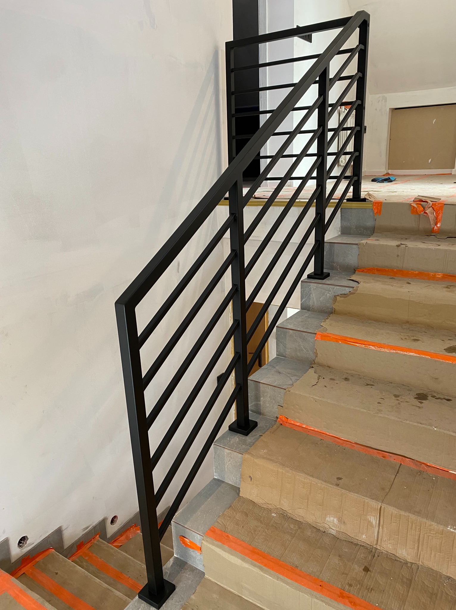 Minimalistyczna, czarna, stalowa balustrada schodowa z poziomymi prętami. Widok z dołu na schody zabezpieczone kartonem. Nowoczesny design.