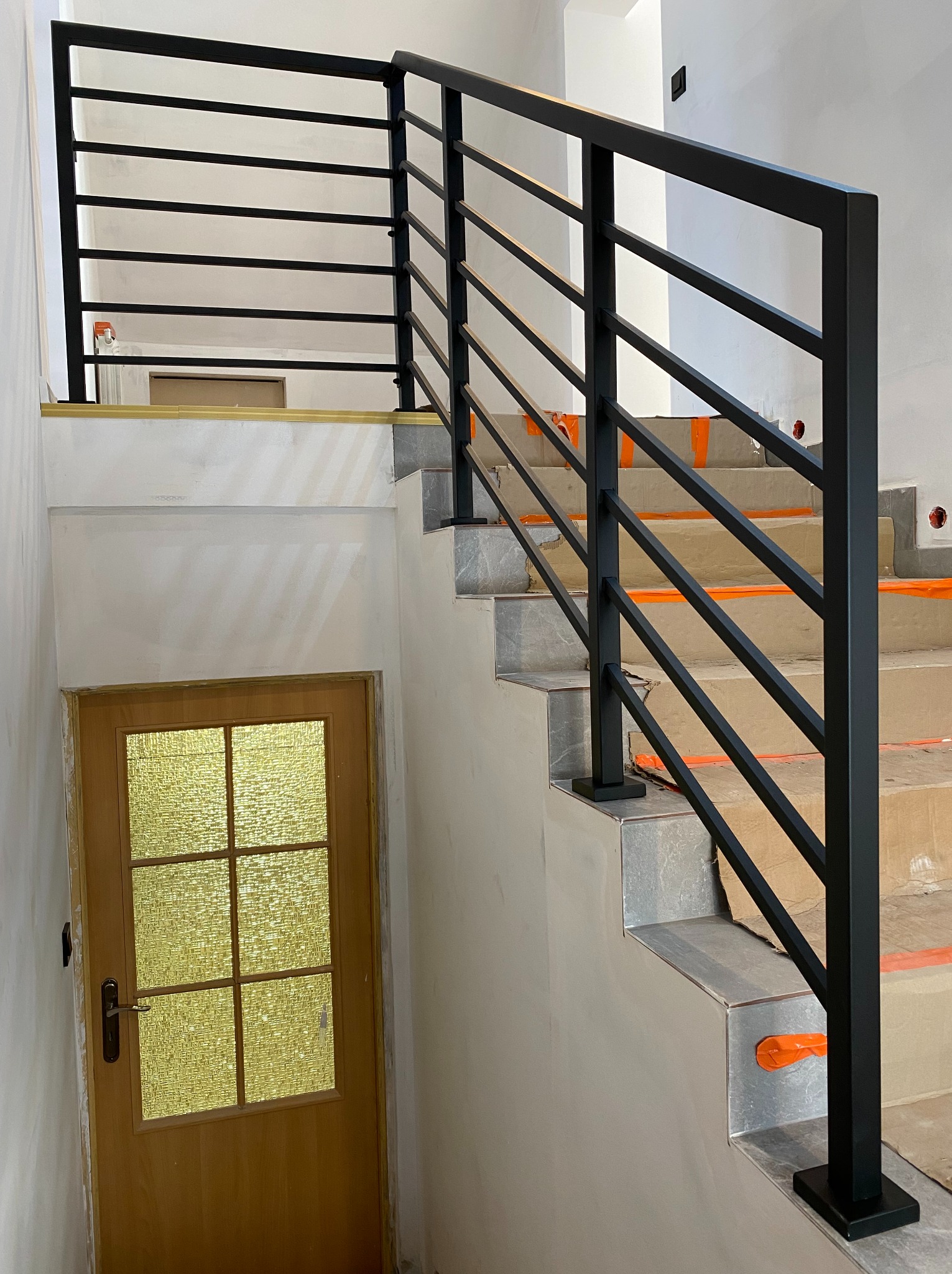Nowoczesna, czarna balustrada metalowa z poziomymi prętami na schodach wykończonych szarą płytką. Widoczna drewniana stolarka drzwiowa z przeszkleniami.