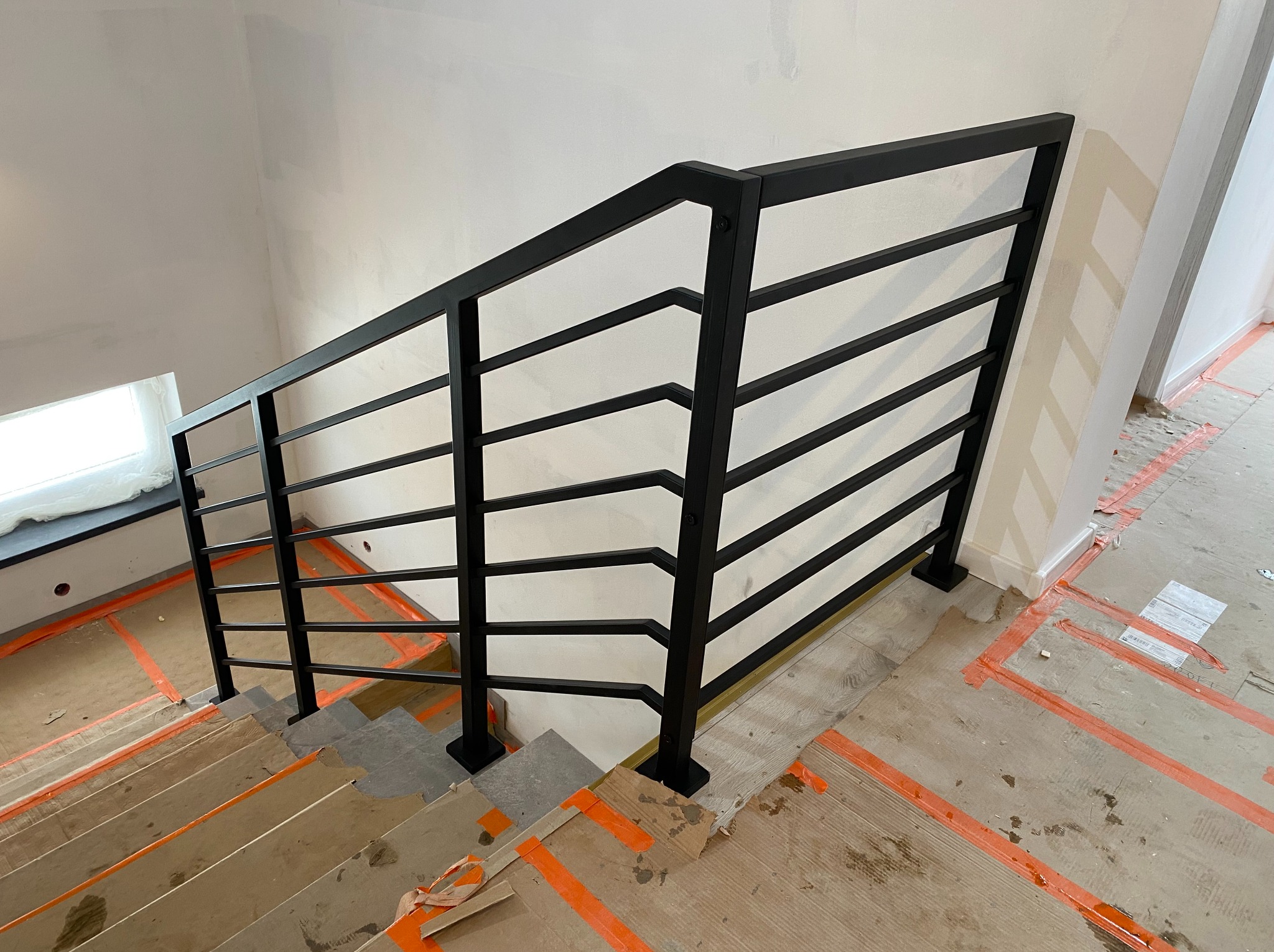 Nowoczesna, czarna balustrada metalowa z poziomymi szczeblami, zamontowana na schodach w trakcie wykończenia. Widoczne zabezpieczenia podłogi i surowe ściany.