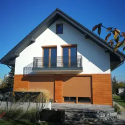 Dwukondygnacyjny dom jednorodzinny z balkonem i drewnianą elewacją na parterze, rolety zewnętrzne opuszczone, widok z ogrodu w słoneczny dzień.