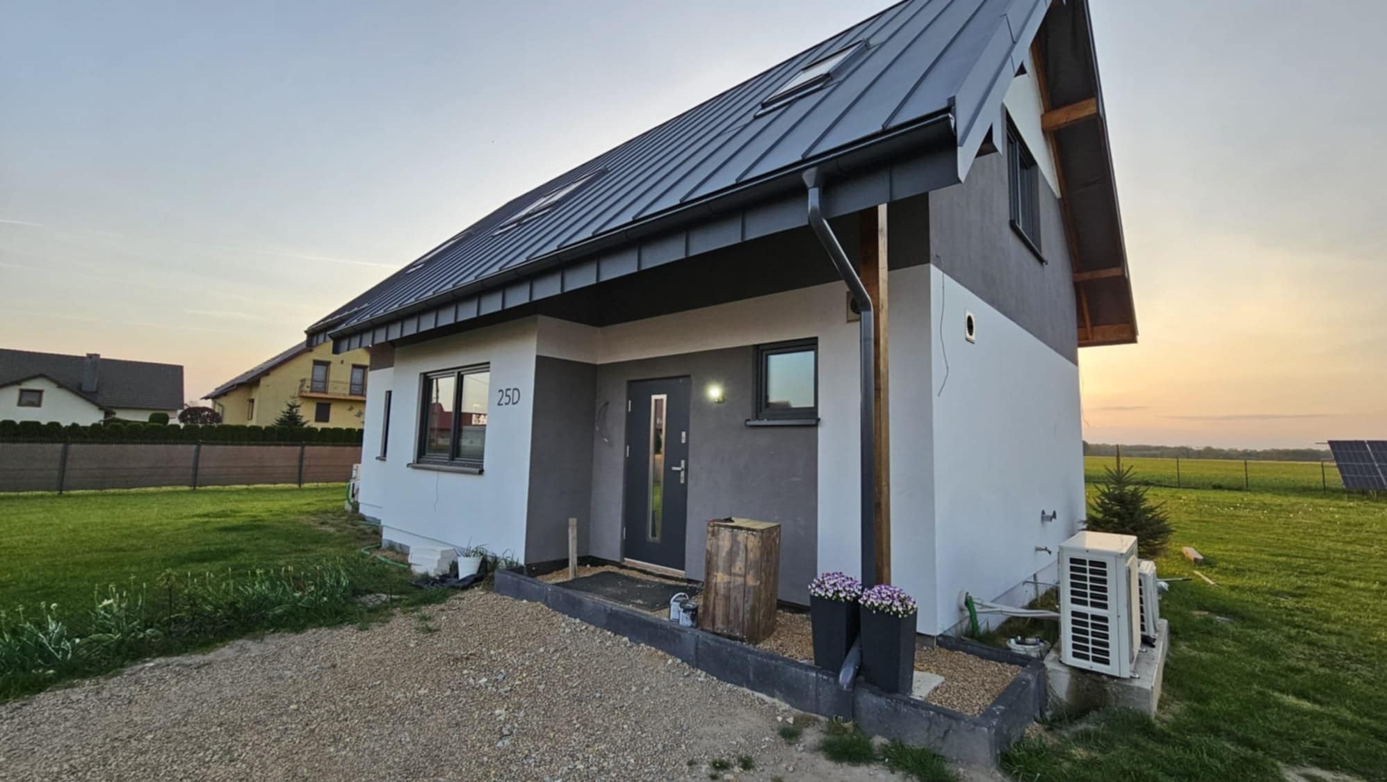 Nowoczesny dom jednorodzinny z dwukolorową elewacją (szary i biały), grafitowym dachem z oknami dachowymi i numerem 25D na ścianie, widok z zewnątrz, w tle panele słoneczne.