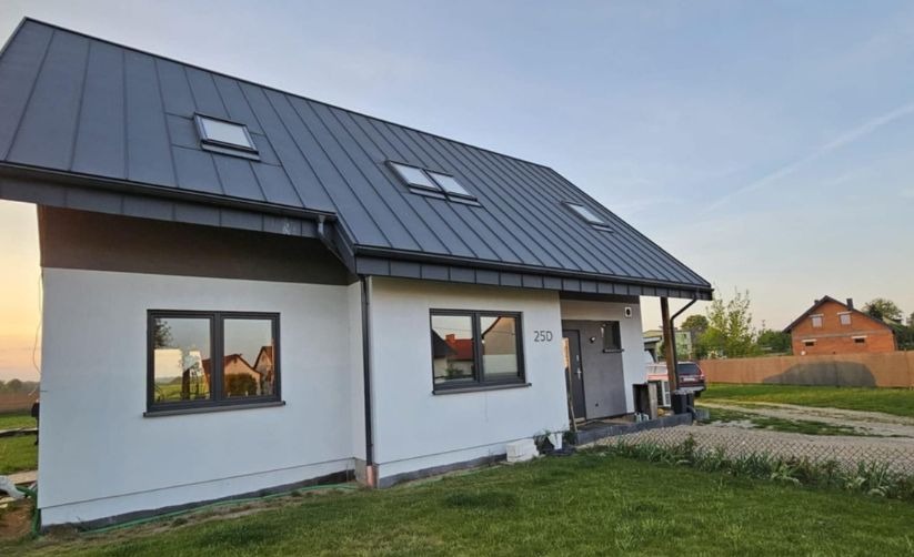 Nowoczesny dom jednorodzinny z szarą blachodachówką i jasnoszarą elewacją, widoczne okna z ciemnymi ramami i numer domu 25D, w tle drugi dom.