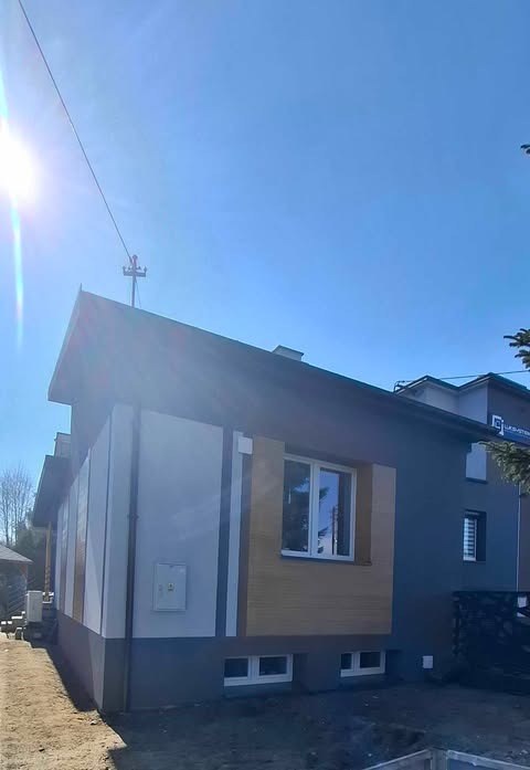 Dom jednorodzinny po częściowej renowacji elewacji: szara ściana z oknem, fragment ocieplony panelami imitującymi drewno, druga ściana biała z szarym cokołem, błękitne niebo.