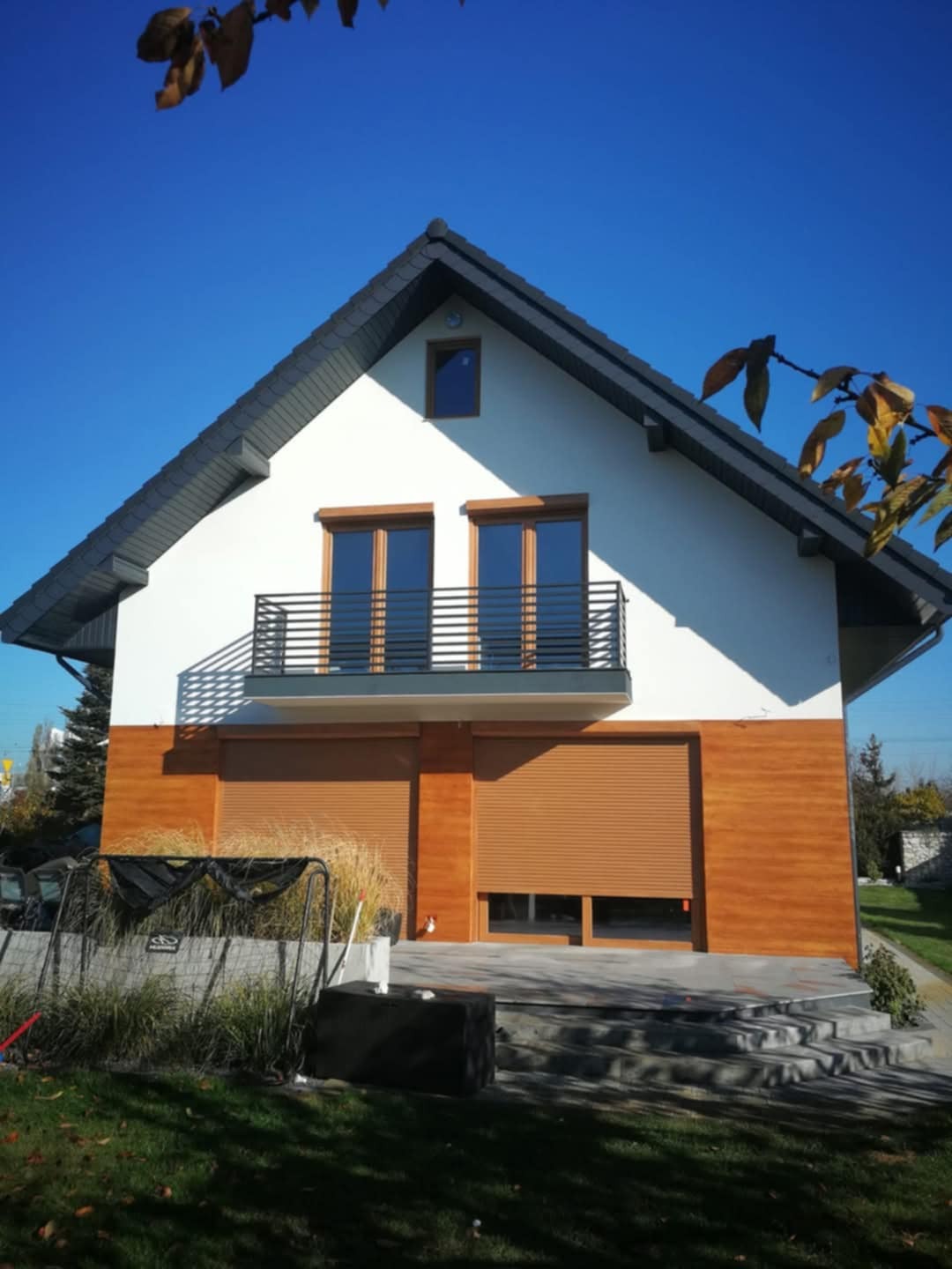 Dwukondygnacyjny dom jednorodzinny z balkonem i drewnianą elewacją na parterze, rolety zewnętrzne opuszczone, widok z ogrodu w słoneczny dzień.