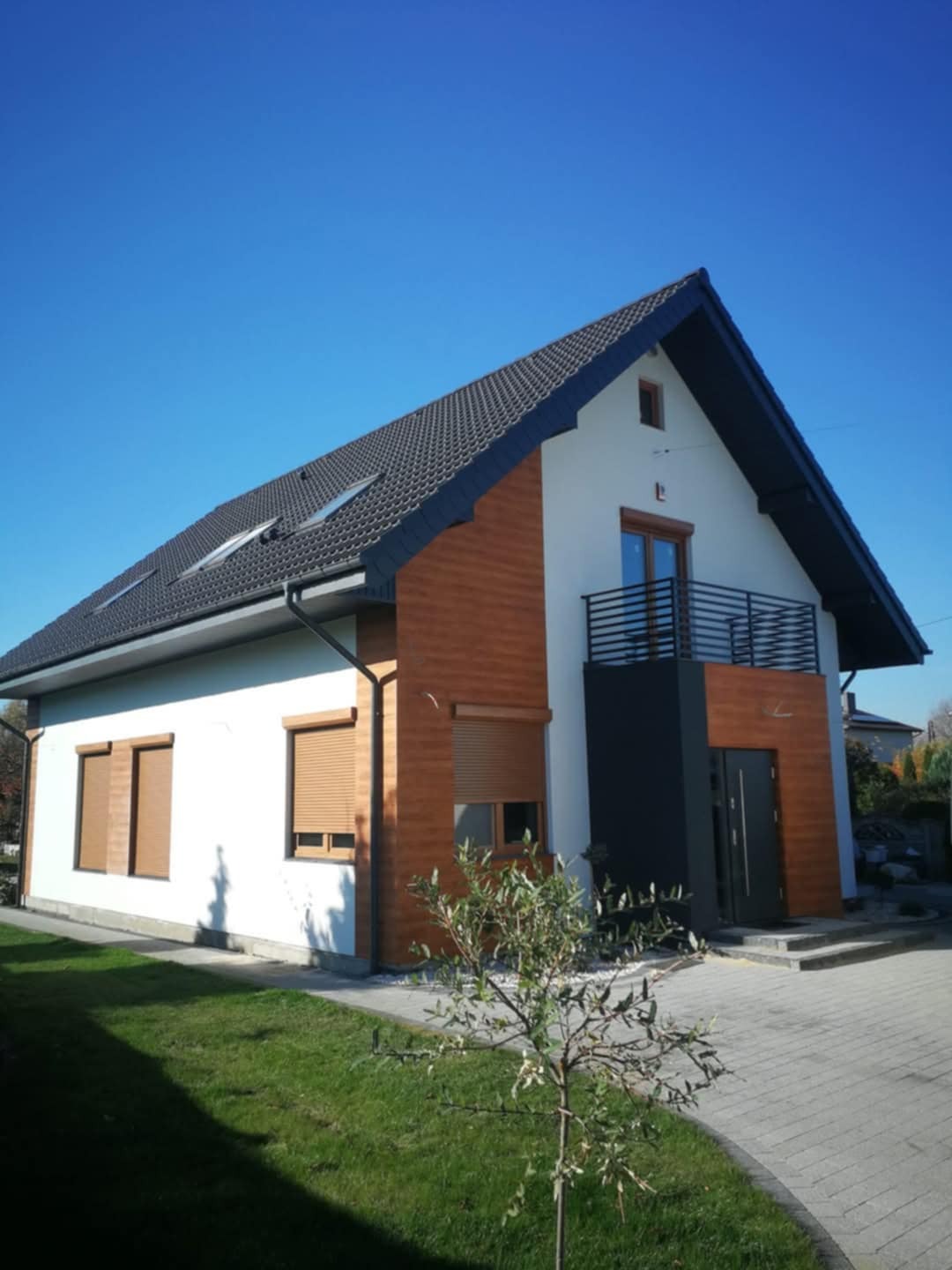 Nowoczesny dom jednorodzinny z elementami drewnopodobnymi na elewacji, ciemną stolarką okienną i drzwiami wejściowymi, oraz zadbanym trawnikiem i młodym drzewkiem na pierwszym planie.