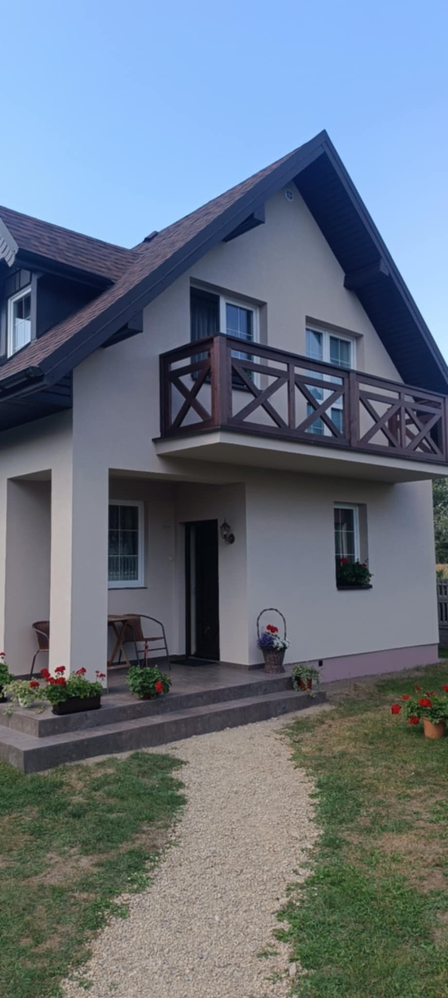 Jasny dom jednorodzinny z ciemnobrązowym dachem i balkonem z drewnianą balustradą w kształcie krzyży, czerwone kwiaty w doniczkach na schodach i w ogrodzie.