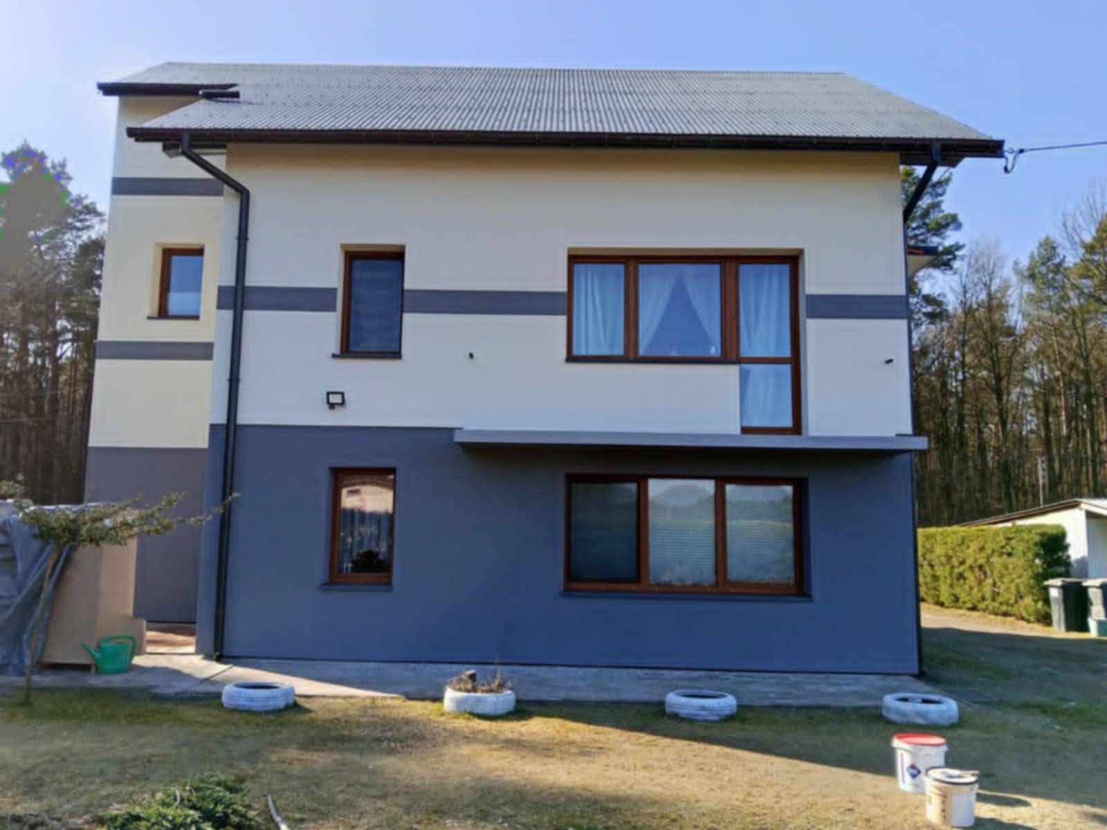 Dwukondygnacyjny dom z elewacją w kolorze szaro-białym, z widocznymi śladami po malowaniu w postaci wiader z farbą i donic z opon na trawniku.