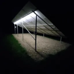 Widok od spodu na panele słoneczne oświetlone nocą, z widoczną konstrukcją wsporczą i podłożem z kamyków.