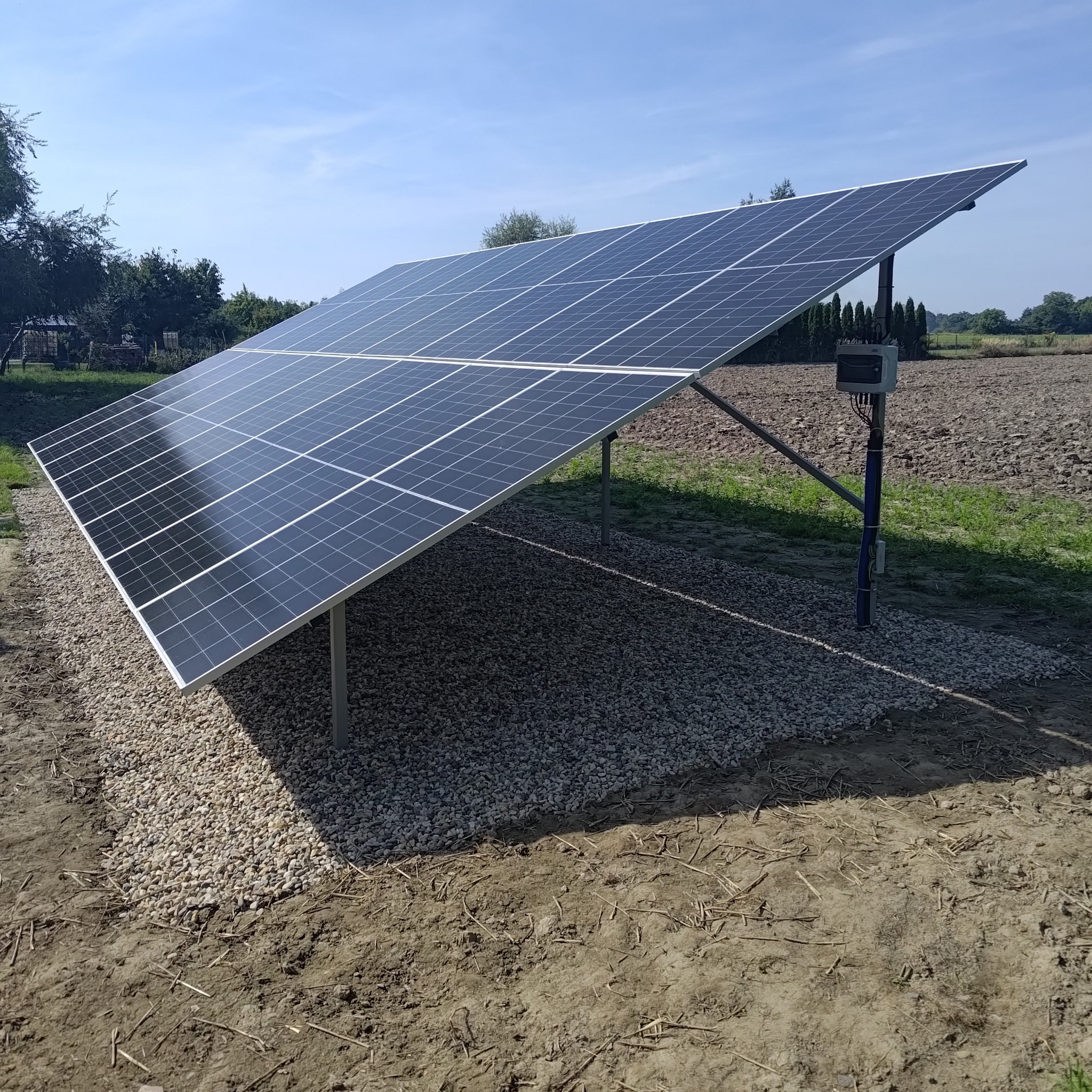 Panel fotowoltaiczny na metalowej konstrukcji, otoczony żwirem, z widoczną skrzynką elektryczną i polami w tle pod błękitnym niebem.