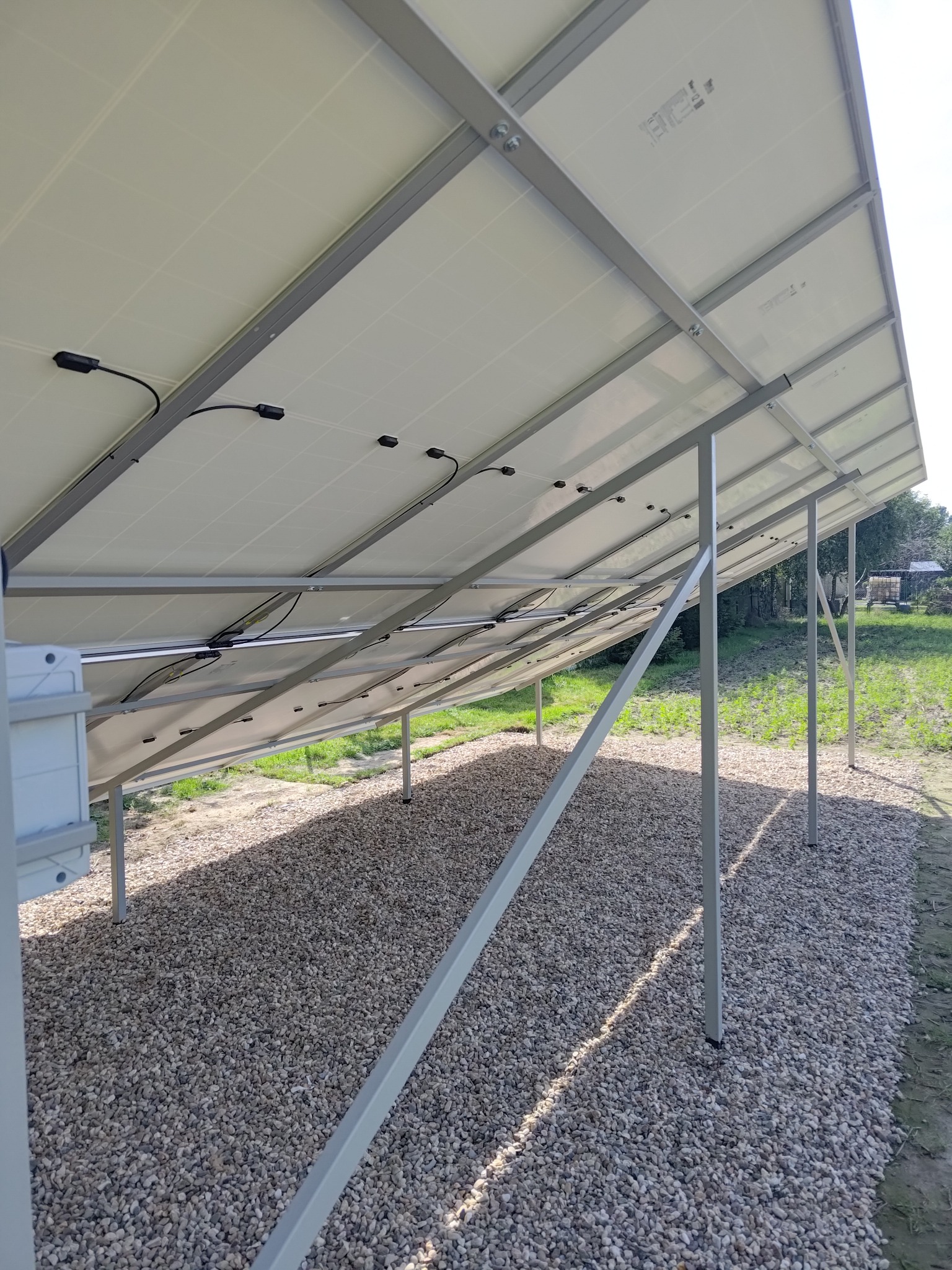 Widok od spodu na konstrukcję wsporczą paneli fotowoltaicznych z widocznymi kablami i elementami montażowymi, posadowioną na żwirowym podłożu z elementami zieleni w tle.