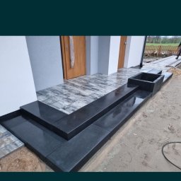 Usługi brukarskie Damian Żuk - Nowoczesne schody wejściowe z czarnej kostki brukowej, prowadzące do domu z szarą elewacją i drewnianymi drzwiami; widoczne także fragmenty chodnika z kostki i rabata kwiatowa w obudowie z kostki.