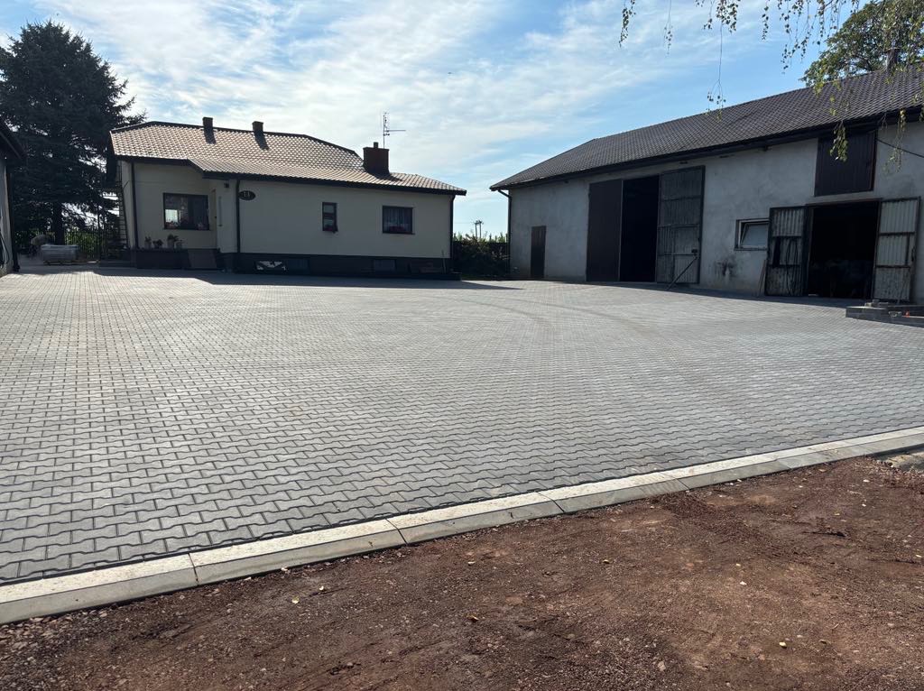 Rozległy, szary plac z kostki brukowej ułożony przed budynkiem mieszkalnym i gospodarczym z otwartymi wrotami, obrzeża z jasnych krawężników, widoczne świeżo obsypane ziemią.