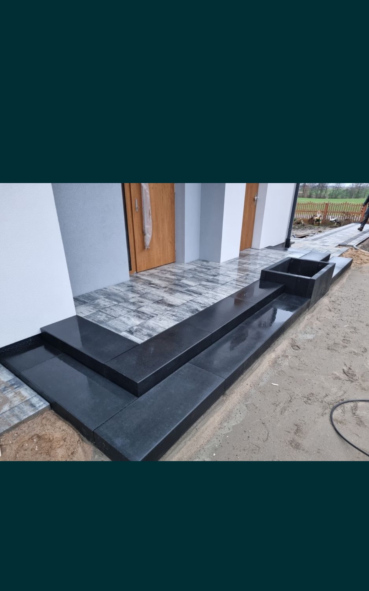 Nowoczesne schody wejściowe z czarnej kostki brukowej, prowadzące do domu z szarą elewacją i drewnianymi drzwiami; widoczne także fragmenty chodnika z kostki i rabata kwiatowa w obudowie z kostki.