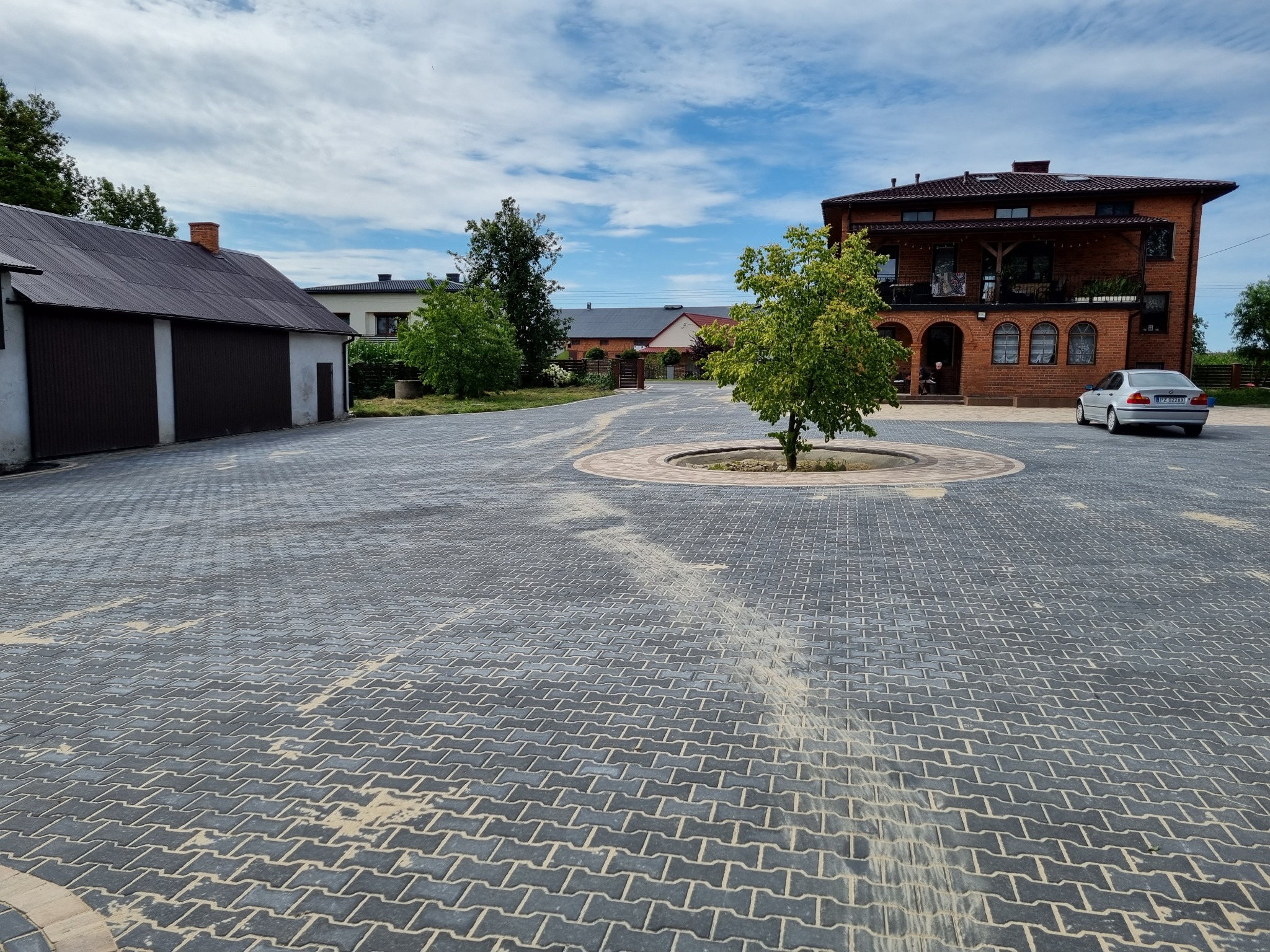 Szeroki plac wyłożony szarą kostką brukową w jodełkę, z okrągłym klombem z drzewem pośrodku, na tle dwupiętrowego domu z czerwonej cegły i zabudowań gospodarczych, częściowo zabrudzony piaskiem.
